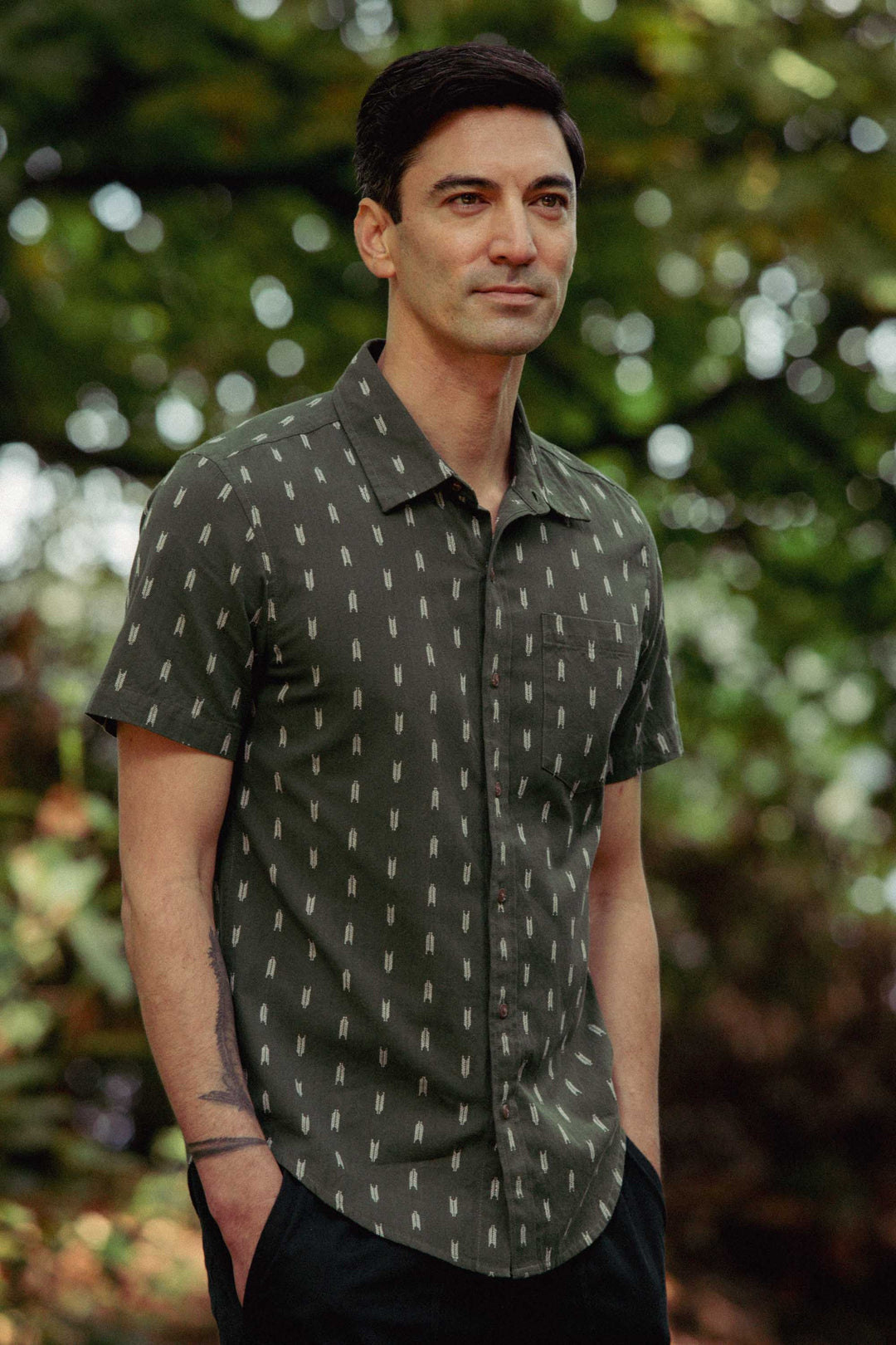 A man with short dark hair wears the Bridge & Burn Harbor Slim Shirt in Thyme Arrows, a lightweight cotton shirt with a subtle pattern. He stands outdoors among trees, looking calm with his hands in his pockets.