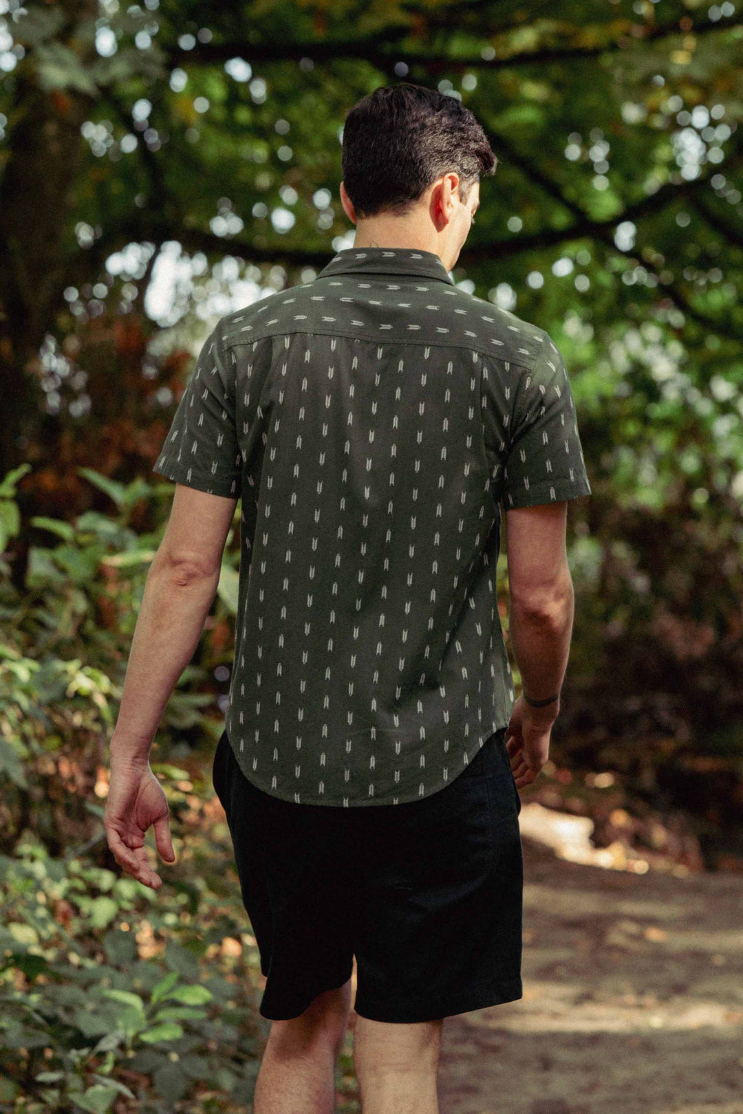 A man with short dark hair walks along a sunlit forest path, wearing the Bridge & Burn Harbor Slim Shirt in Thyme Arrows and black shorts. His back faces the camera, surrounded by lush greenery.