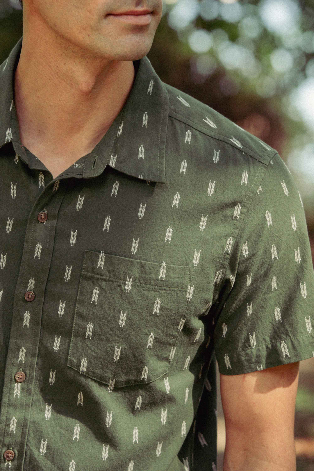 Wearing the Bridge & Burn Harbor Slim Shirt in Thyme Arrows, a person stands outdoors; only the lower half of their face and upper torso are visible.