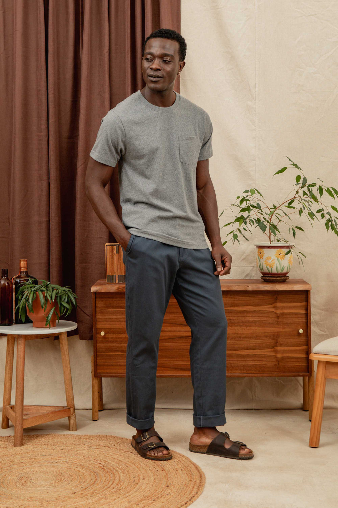 A man stands indoors, smiling with his hands in his pockets. He's wearing Bridge & Burn's Hayes Pant in Dark Slate, a gray t-shirt, and brown sandals. Behind him are a wooden console table, plant, and round rug.