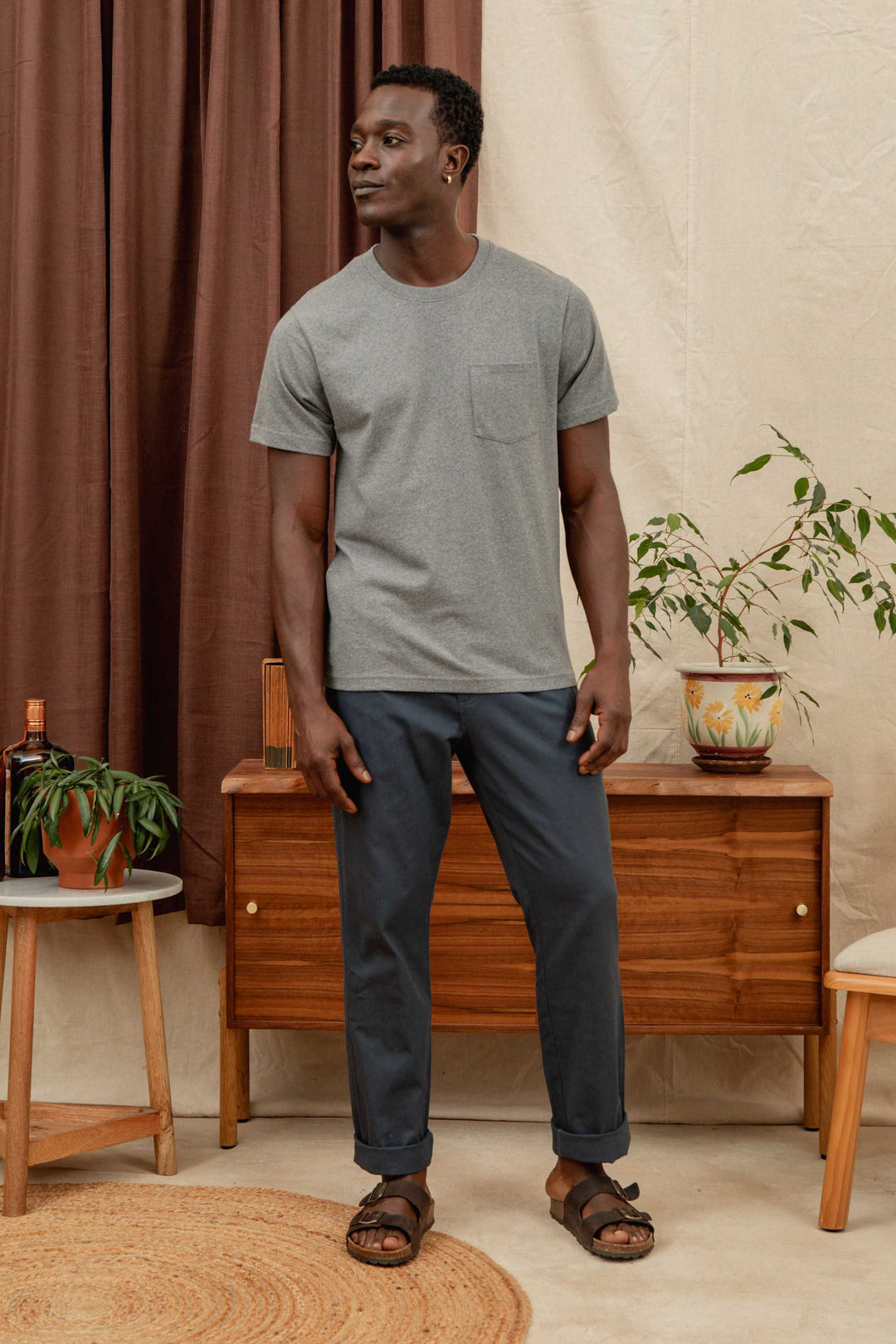 A man wearing a gray t-shirt, Bridge & Burn Hayes Pant in Dark Slate, and brown sandals stands in a room with mid-century modern furniture, two potted plants, and brown curtains. He looks slightly to the side with a relaxed expression.