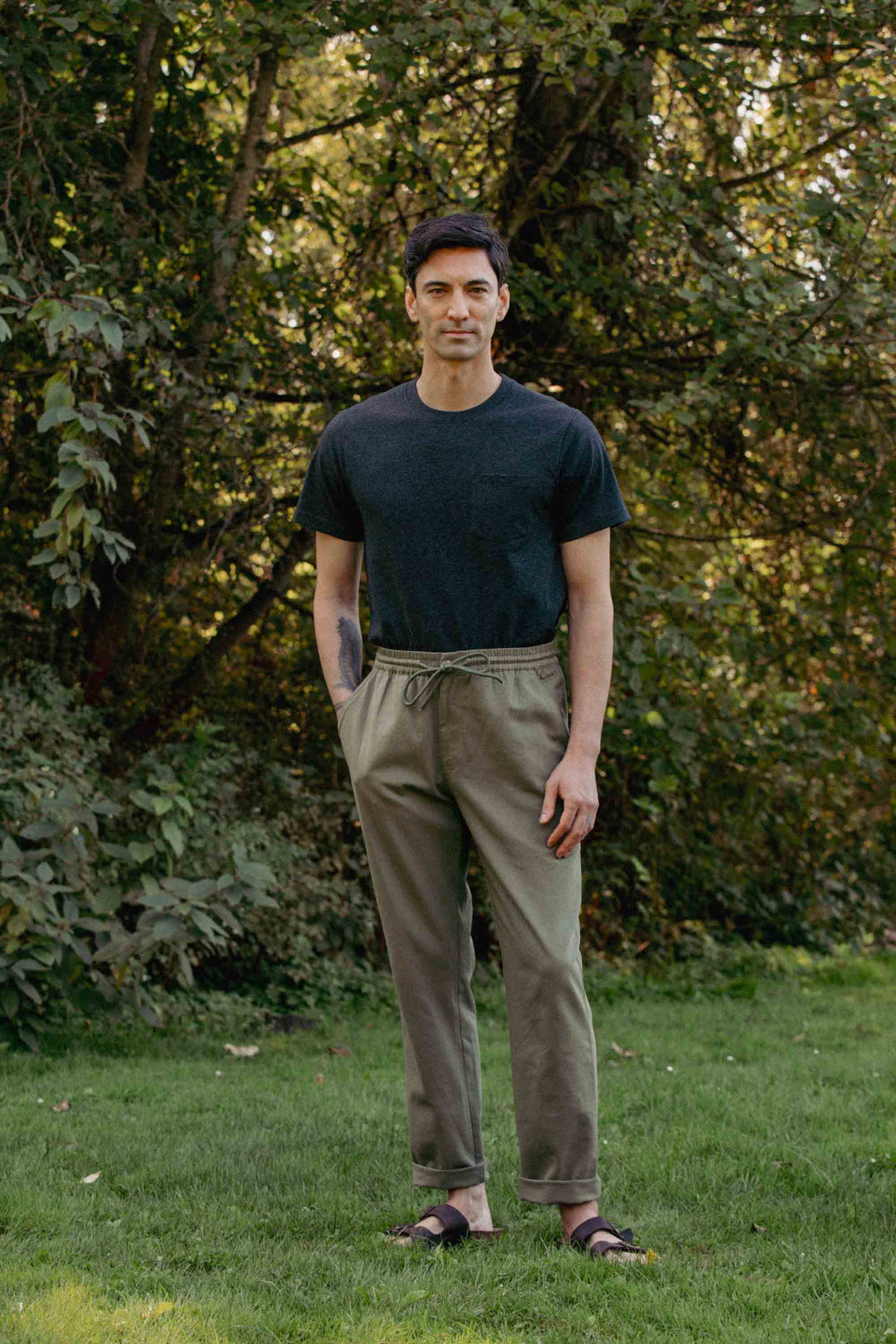 A man stands outdoors on grass in front of leafy trees, wearing Bridge & Burn's Hayes Pant in Olive with an elastic waistband and relaxed fit, paired with a dark T-shirt and sandals. His hands are in his pockets as he faces the camera casually.