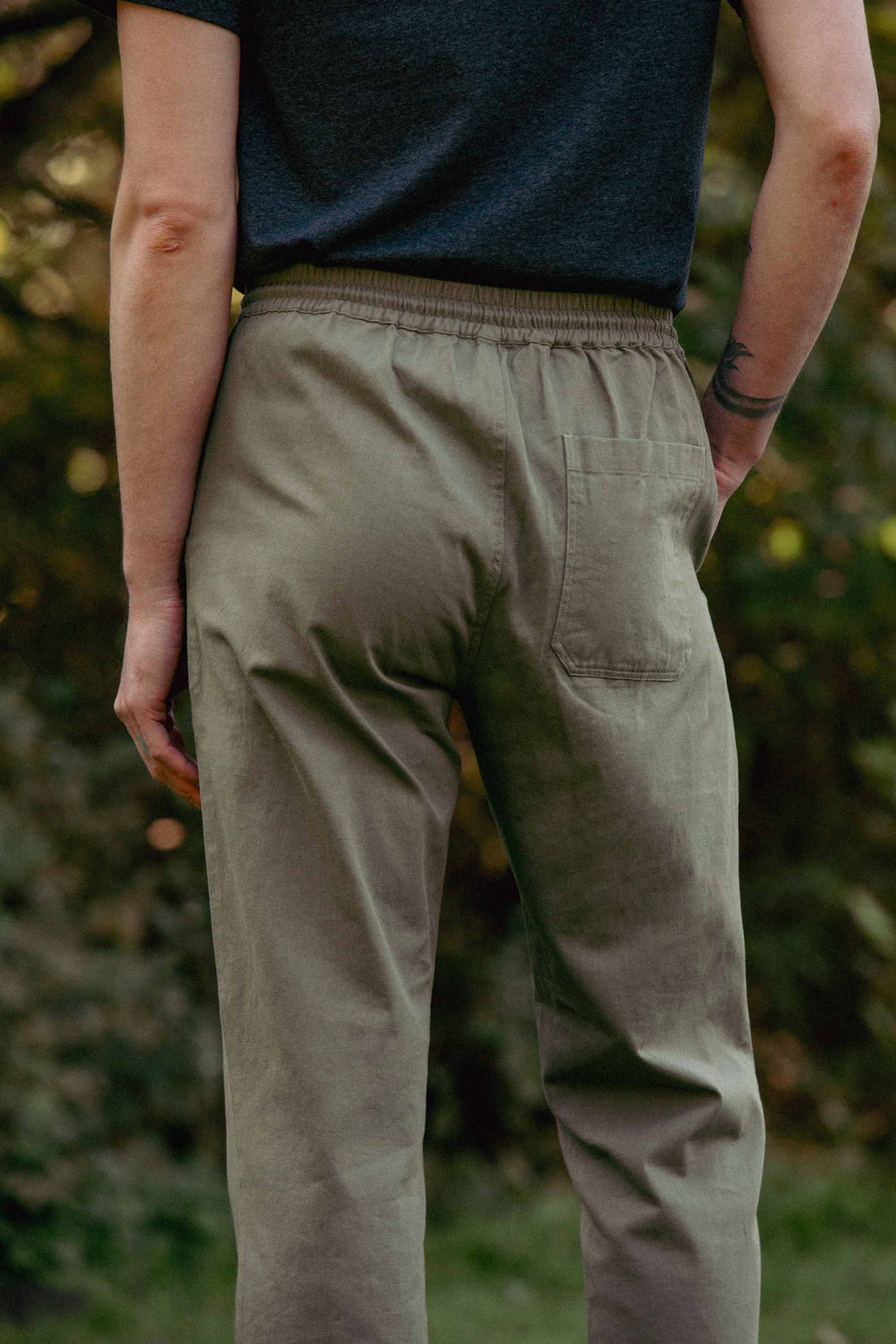 A person stands outdoors, facing away from the camera, wearing Bridge & Burn's Hayes Pant in olive. The relaxed fit pants feature an elastic waistband and back pocket. Their tattooed left arm is visible against a backdrop of blurred greenery.