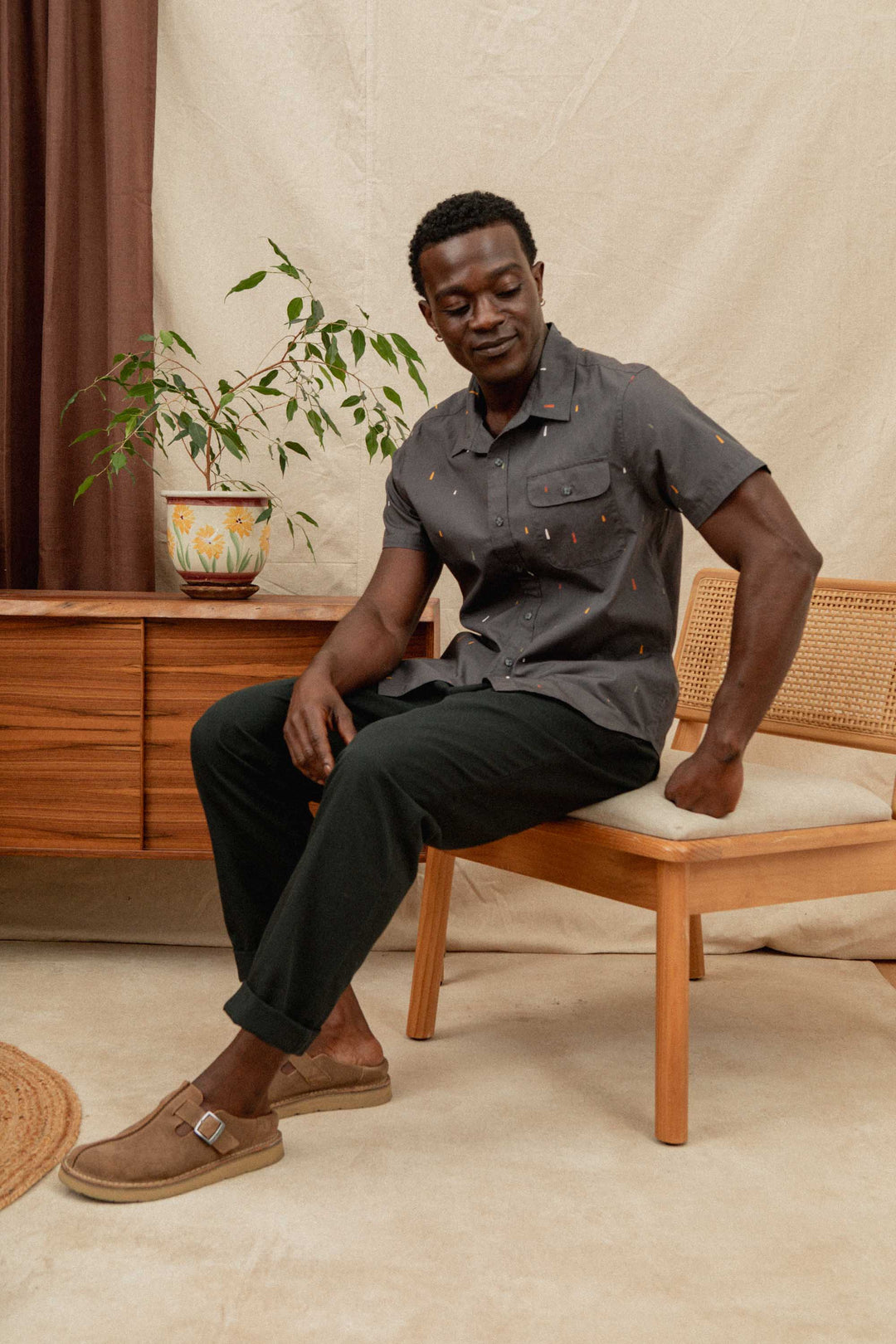 A man wearing a short-sleeve gray shirt and Bridge & Burn Hayes Pant in Soft Black sits on a wooden chair, looking down with one hand on his knee. He wears brown shoes; a sideboard and potted plant are behind him.
