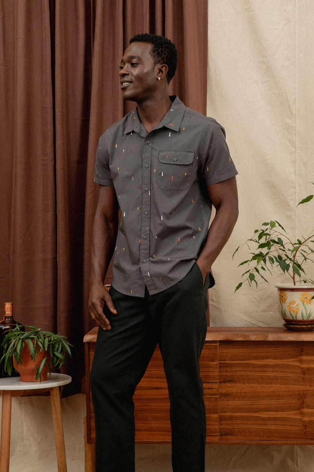 A man stands indoors, smiling with one hand in his pocket, wearing relaxed Bridge & Burn Hayes Pant in Soft Black and a short-sleeved gray button-up shirt. Brown curtains, wooden furniture, and potted plants are behind him.