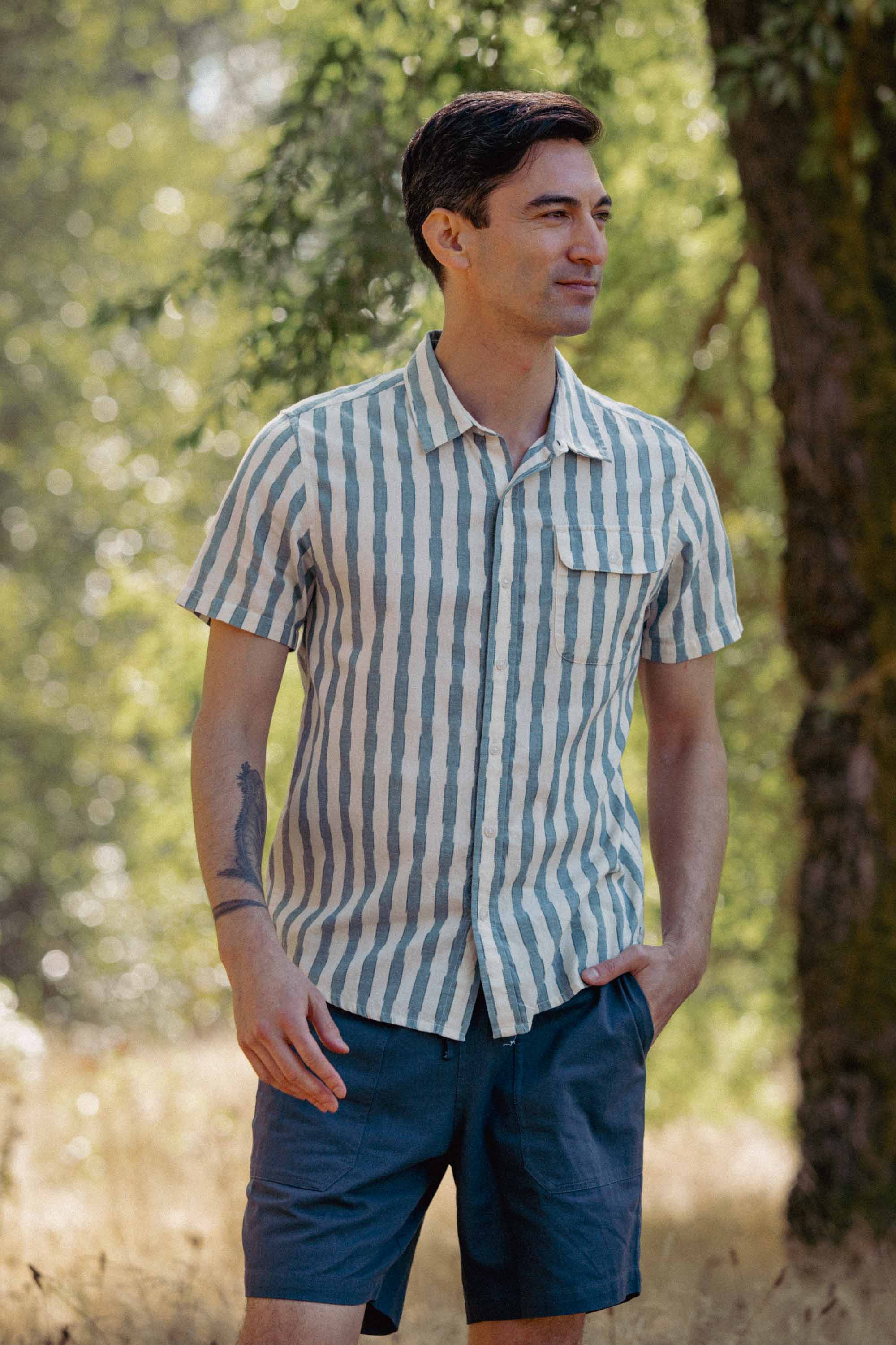 A man stands outdoors in sunlight, wearing the Bridge & Burn Marten Shirt in Beach Stripe with blue shorts. Surrounded by trees, he has one hand in his pocket and a tattoo on his left forearm.