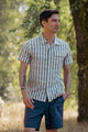 A man stands outdoors in sunlight, wearing the Bridge & Burn Marten Shirt in Beach Stripe with blue shorts. Surrounded by trees, he has one hand in his pocket and a tattoo on his left forearm.