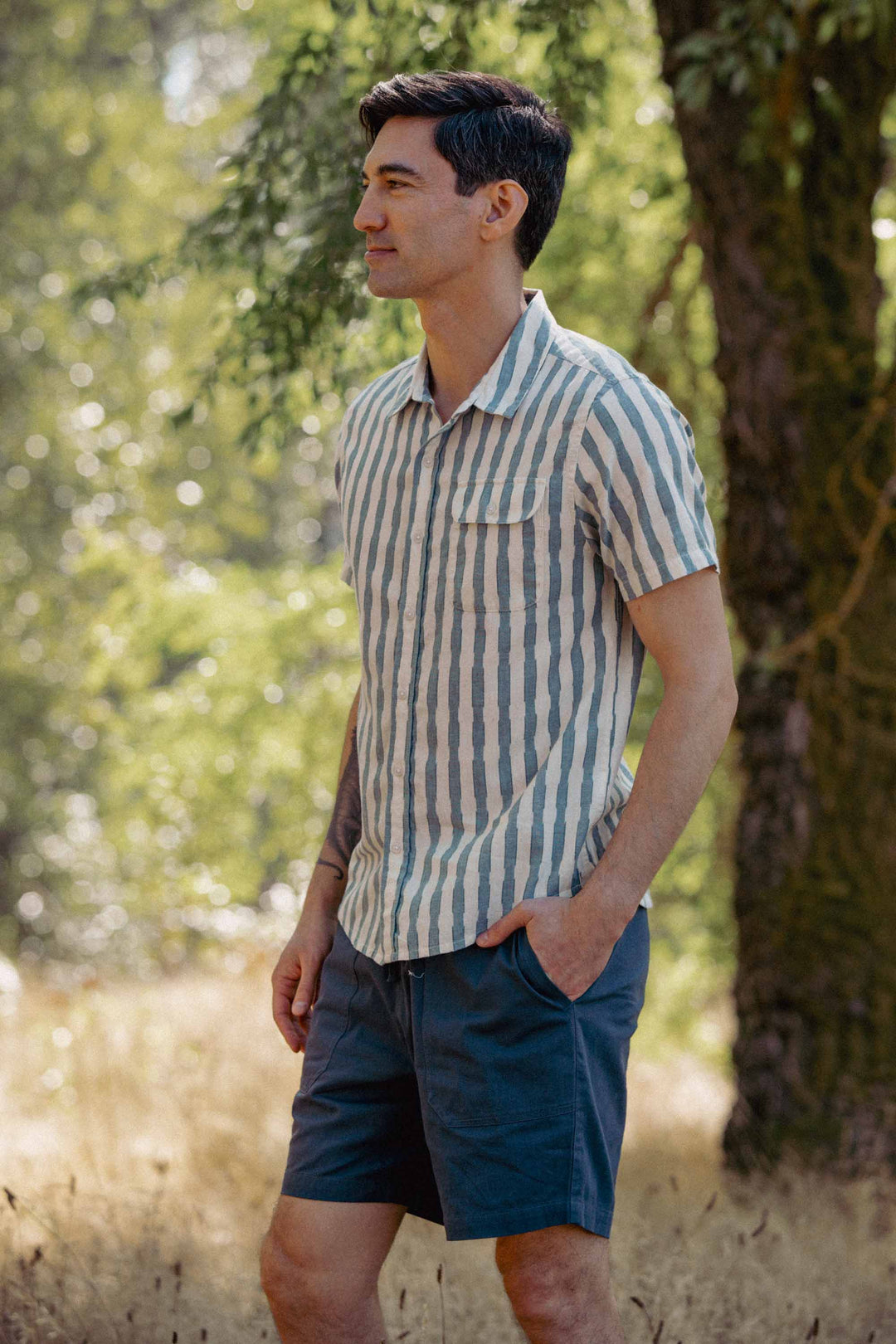 A man stands outdoors in a grassy area with trees, wearing the Bridge & Burn Marten Shirt in Beach Stripe and blue shorts. His hands are in his pockets and he looks to the side, smiling slightly.