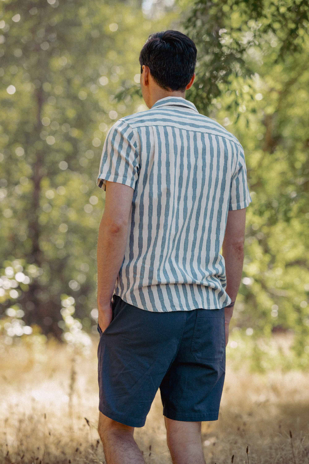 A person stands outdoors with their back to the camera, wearing the Bridge & Burn Marten Shirt in Beach Stripe and dark blue shorts. Their hands are in their pockets as trees and sunlight fill the background.
