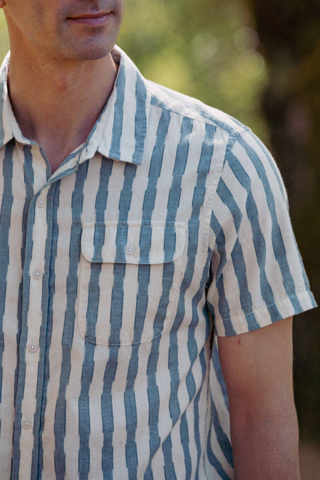 A man stands outdoors, sunlight softly illuminating the Bridge & Burn Marten Shirt in Beach Stripe—a cotton casual shirt featuring classic-fit vertical blue and white stripes. Only his upper body and part of his face are visible.