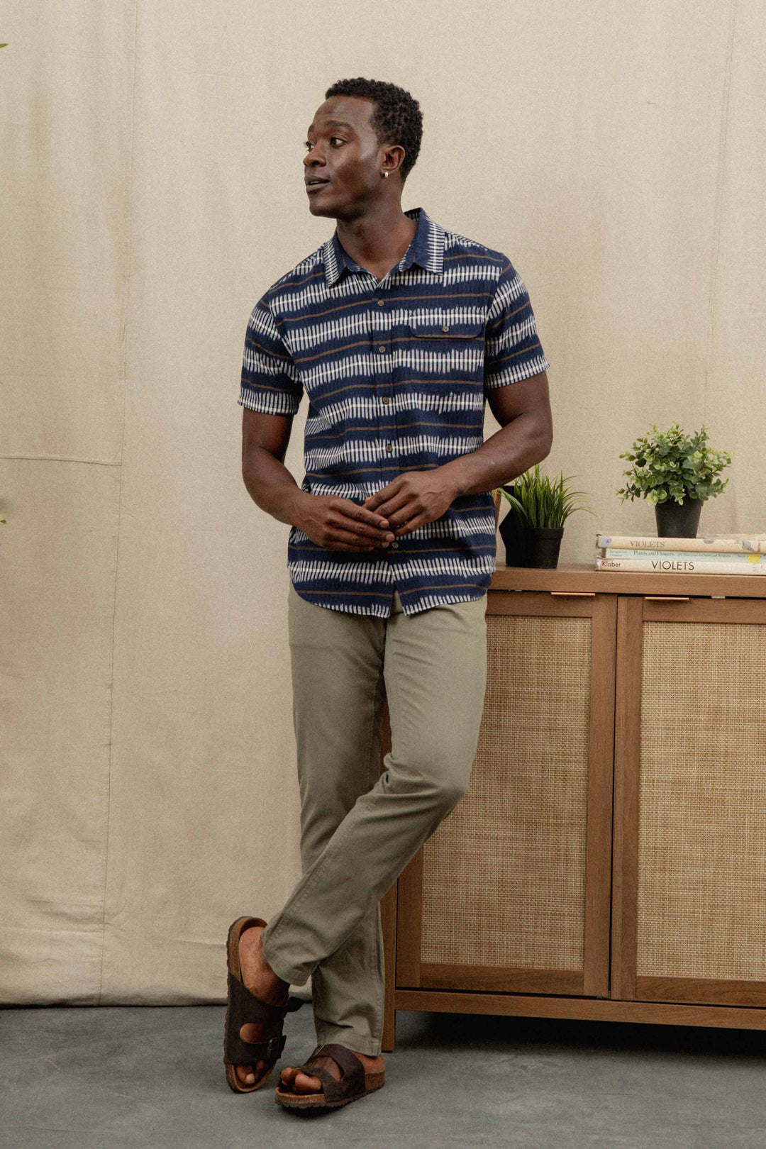 A man wearing the Bridge & Burn Marten Shirt in Dusk Mirage, khaki pants, and sandals leans against a wooden cabinet with potted plants and books, set before a beige curtain backdrop.