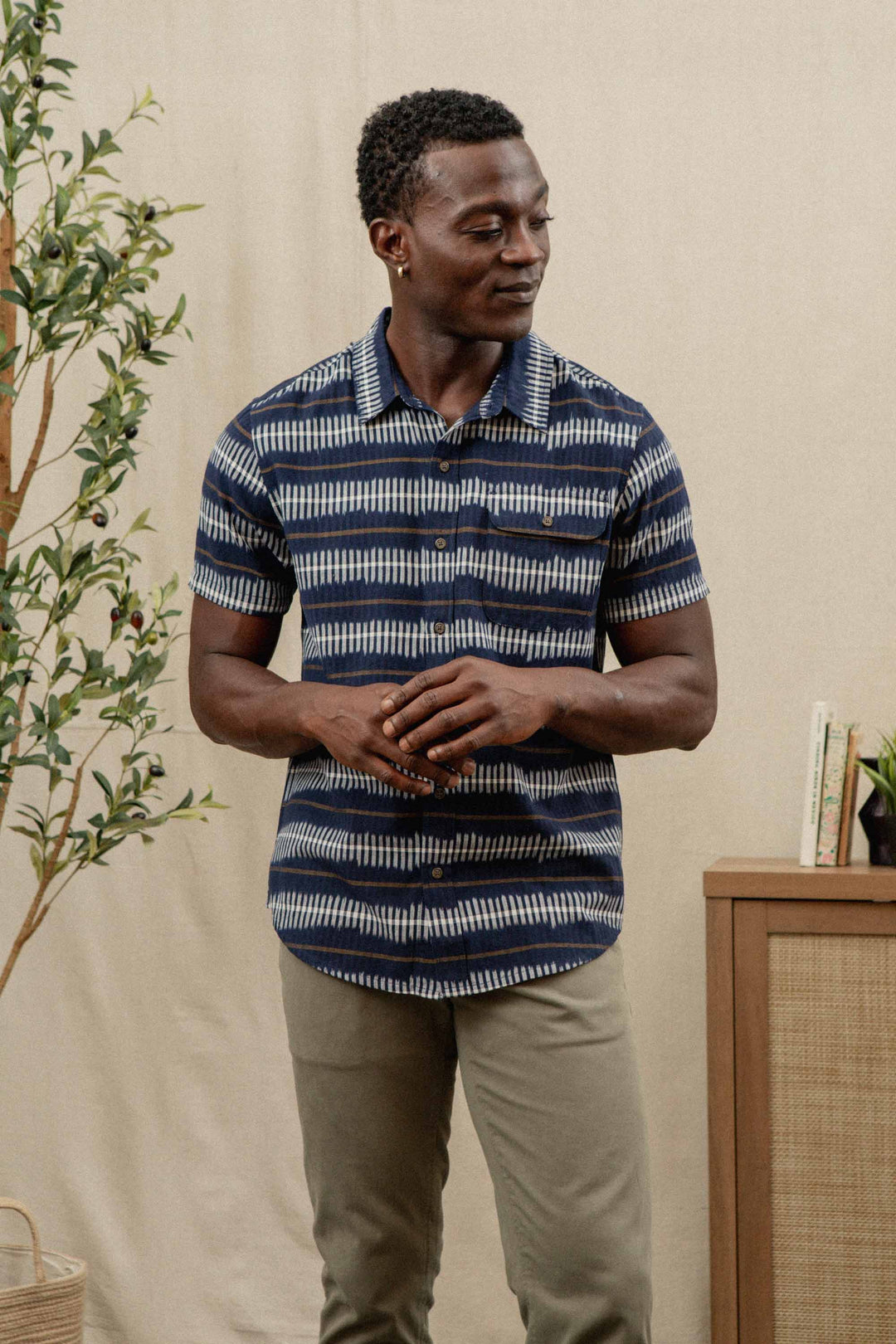 A man stands indoors wearing the Bridge & Burn Marten Shirt in Dusk Mirage, a navy blue and white patterned cotton short-sleeve shirt, paired with light khaki pants. He looks to the side with hands clasped, a plant and books on the cabinet behind him.