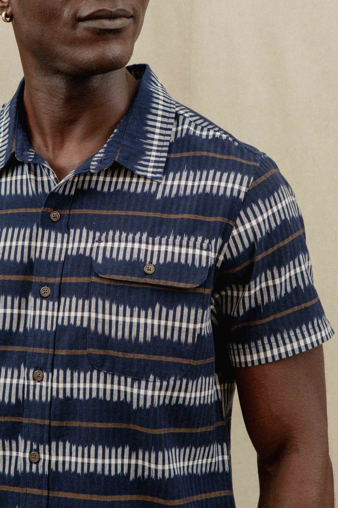 A man wears the Bridge & Burn Marten Shirt in Dusk Mirage—a classic fit, short sleeve button-down with navy, white, and brown stripes—standing against a neutral background. Only his upper body and part of his face are visible.