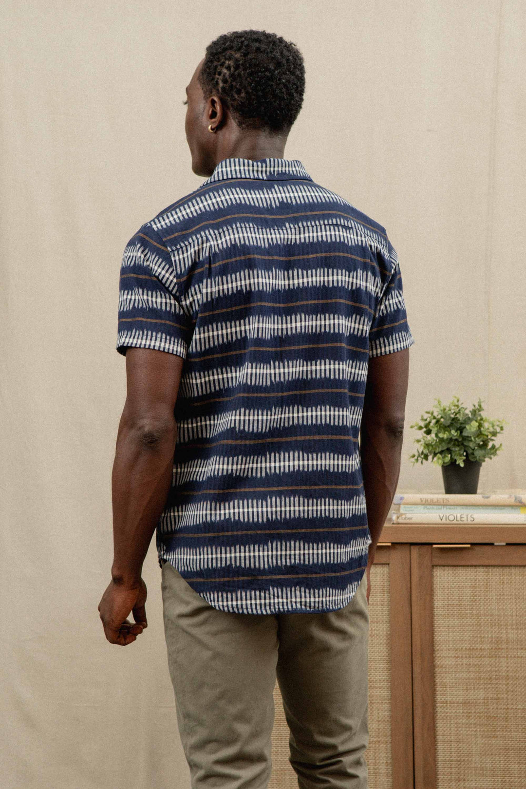 A man faces away, wearing the Bridge & Burn Marten Shirt in Dusk Mirage, a blue and white striped cotton short sleeve button-down, paired with khaki pants. In the background are a small plant and stacked books on a cabinet.