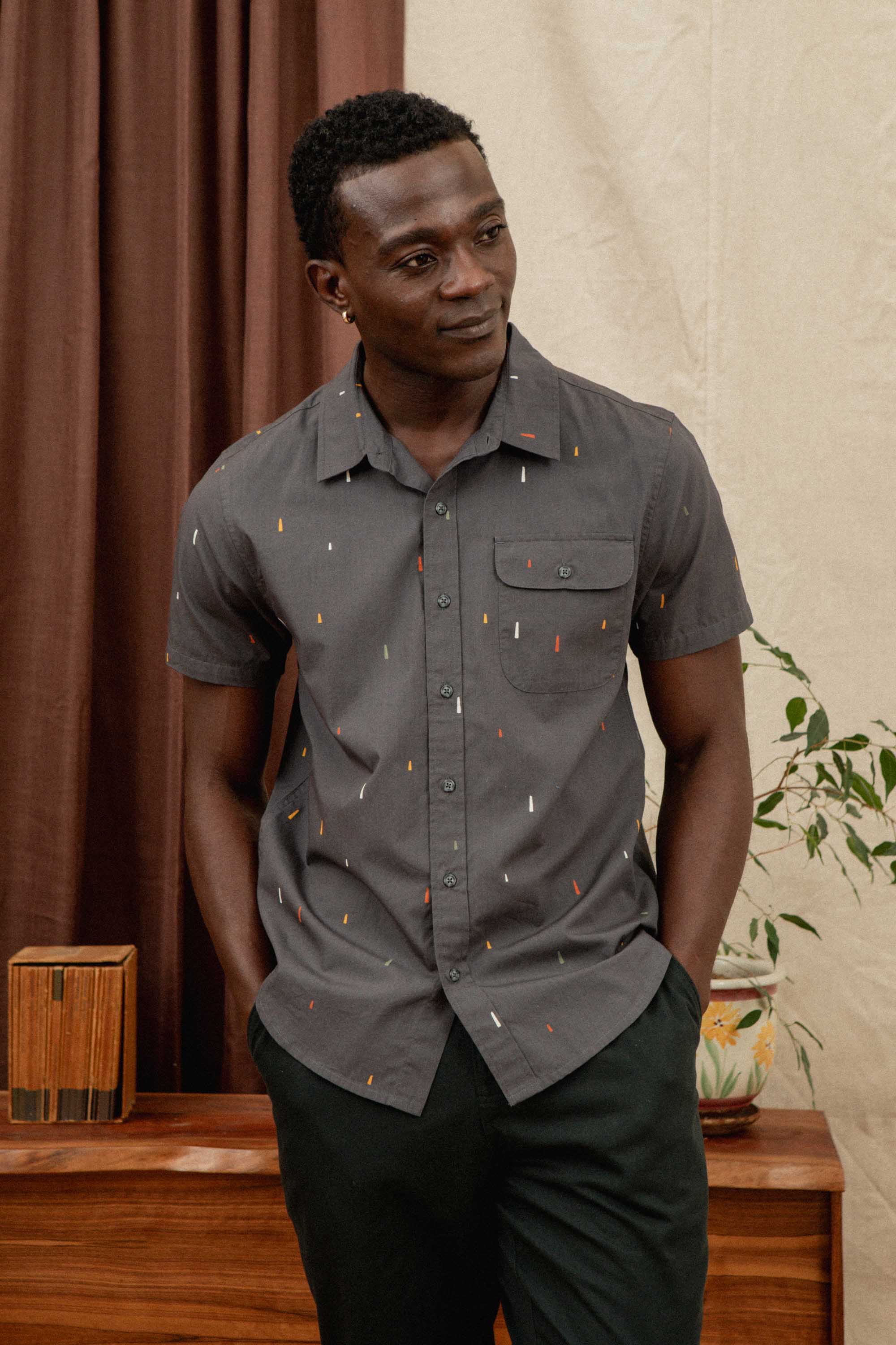 A man wears the Bridge & Burn Marten Shirt in Midnight Rain, a short sleeve button-down with a subtle pattern, standing indoors with hands in pockets near a wooden cabinet, potted plant, and brown curtains.