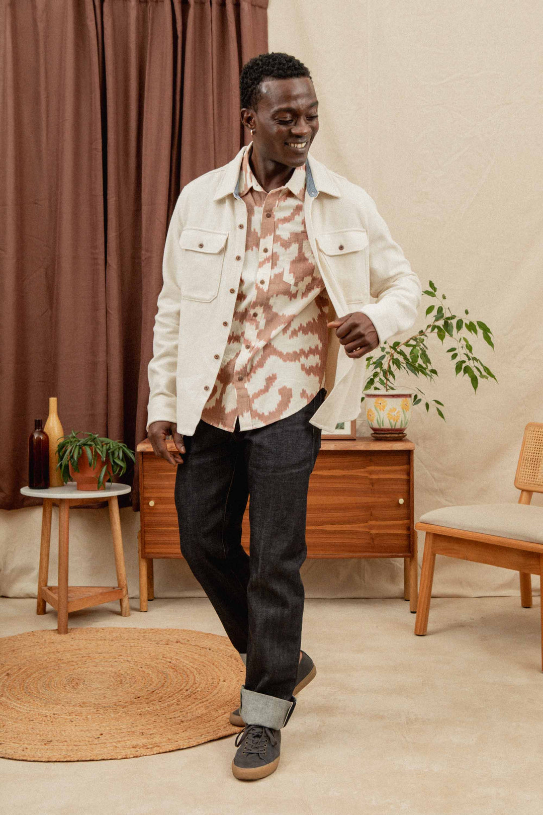 A man wears the Bridge & Burn Pointer Overshirt in Ivory Twill over a patterned shirt, paired with dark cuffed jeans and sneakers. He stands indoors, smiling amid plants, wooden furniture, and a brown curtain in the background.