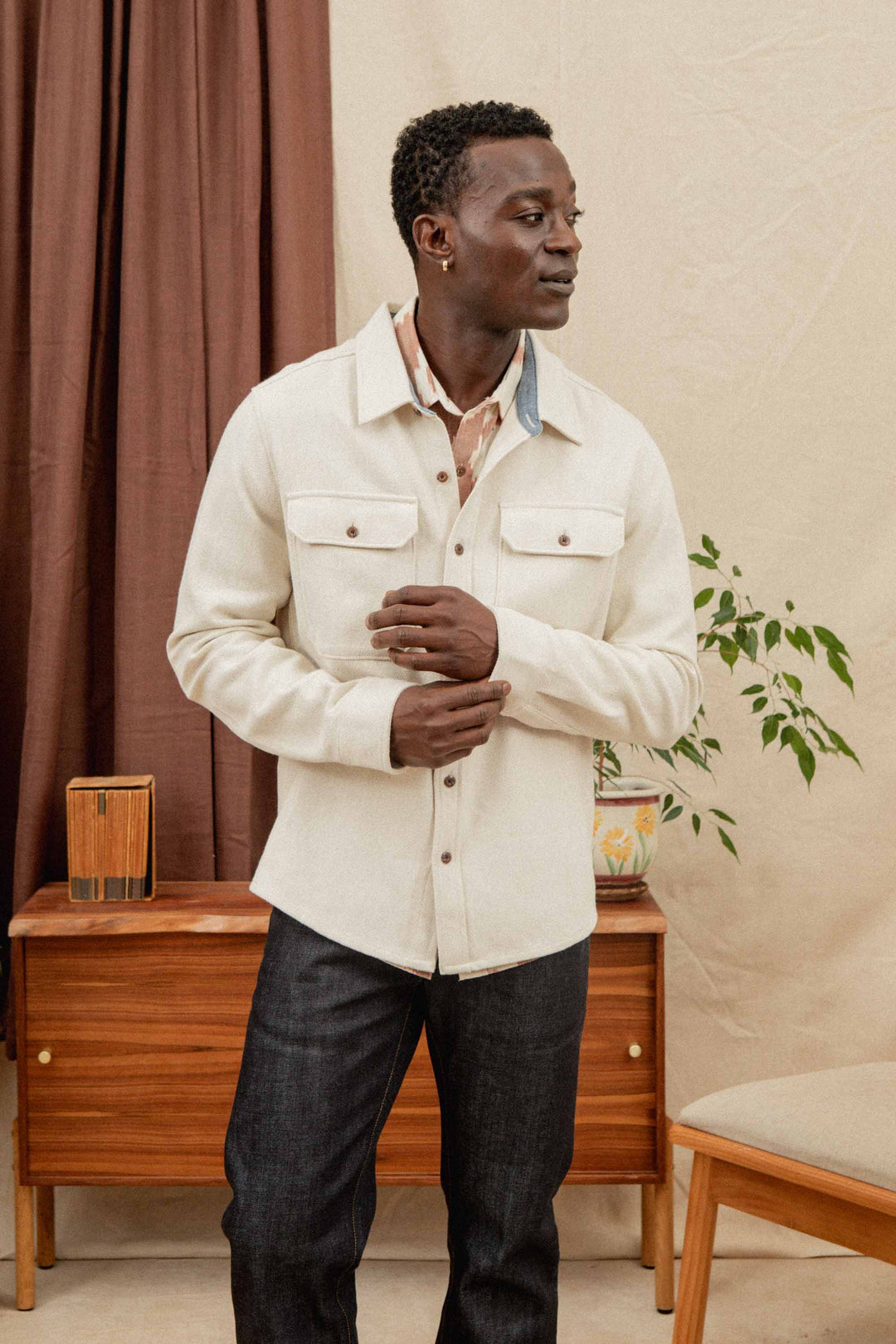 A man wearing the Bridge & Burn Pointer Overshirt in Ivory Twill and dark jeans stands indoors near a wooden cabinet with plants, looking to his left with hands clasped. The background features a brown curtain and beige wall.