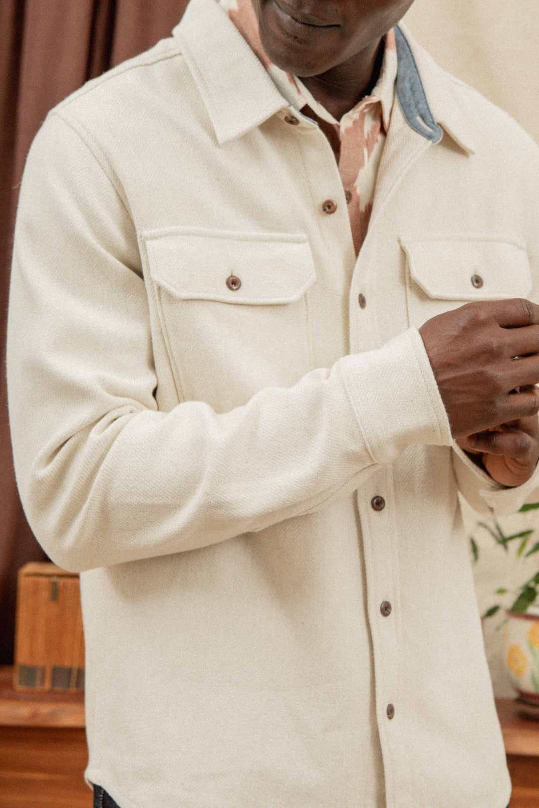 A man in the Bridge & Burn Pointer Overshirt in Ivory Twill—featuring two chest pockets, brown buttons, and crafted from recycled fibers—stands indoors adjusting his sleeve, with part of his face out of frame.