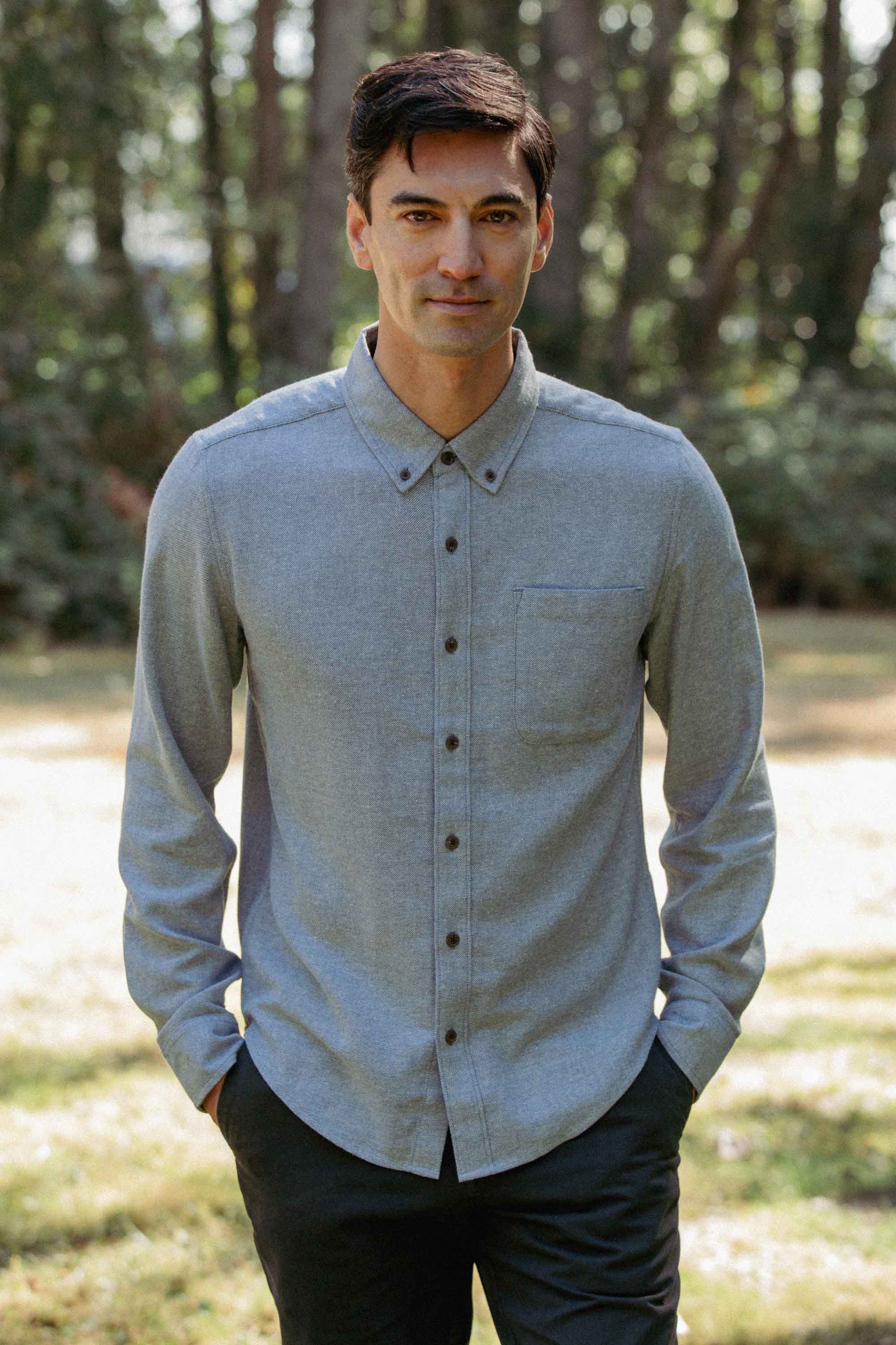 A man stands outdoors with hands in pockets, wearing the Bridge & Burn Sutton Slim Shirt in Denim Twill. He faces the camera, surrounded by blurred trees and sunlight in the background.
