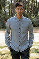 A man stands outdoors with hands in pockets, wearing the Bridge & Burn Sutton Slim Shirt in Denim Twill. He faces the camera, surrounded by blurred trees and sunlight in the background.
