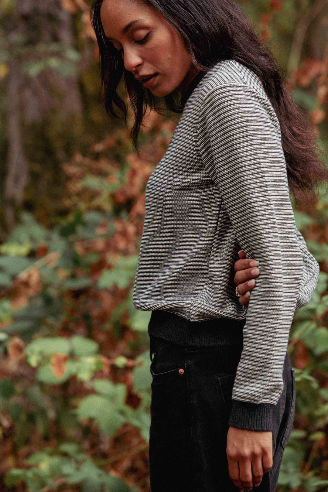A woman with long dark hair stands outdoors among green foliage and a softly blurred background, wearing Bridge & Burn's Ansel Sweater in Charcoal Stripe—an essential mid-weight sweater crafted from recycled fibers—and black pants, looking down with one hand at her side.