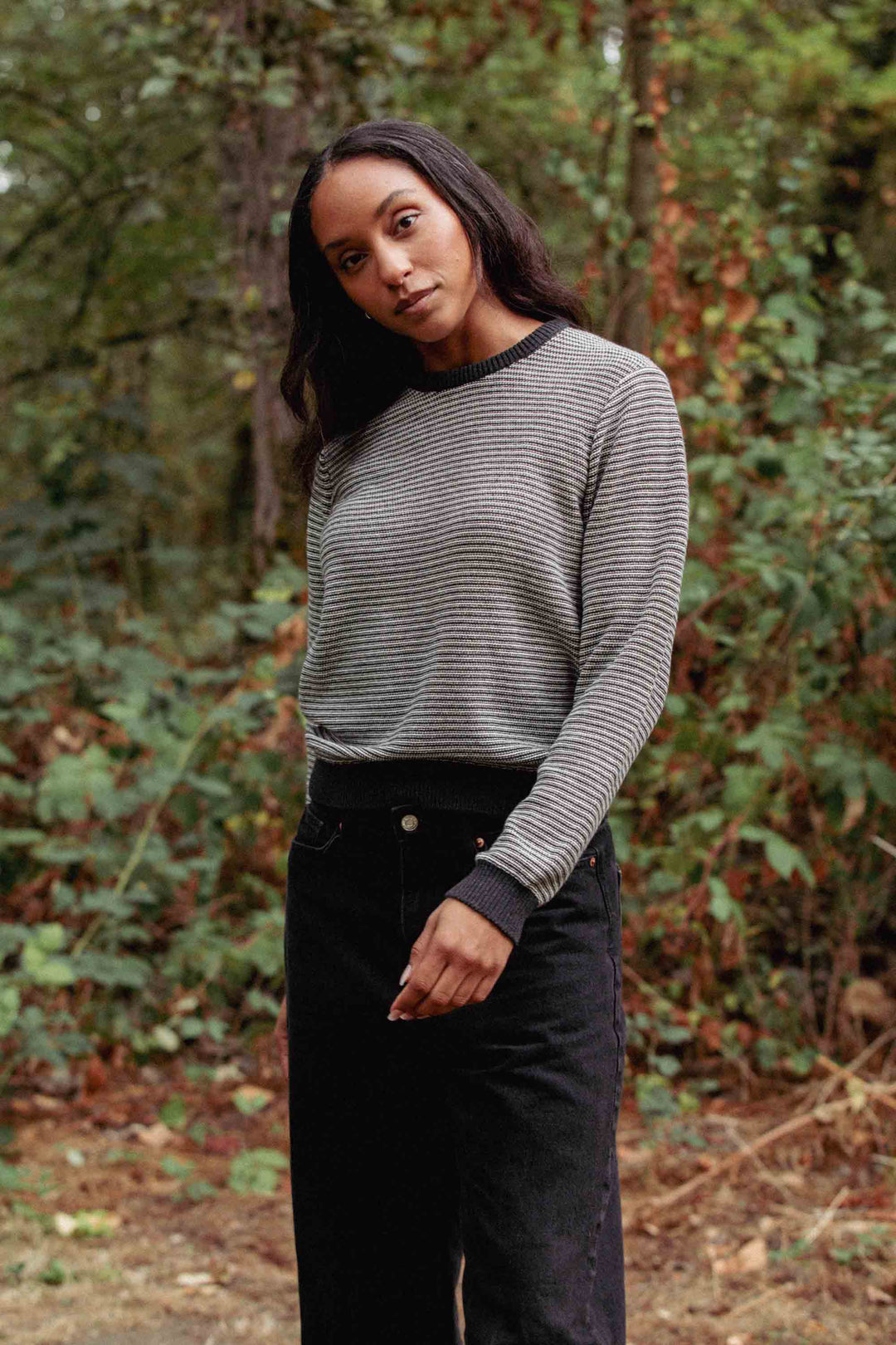 A woman with long dark hair stands outdoors in front of greenery, wearing the Bridge & Burn Ansel Sweater in Charcoal Stripe—a mid-weight sweater made from recycled fibers—paired with black pants, her head tilted slightly and one arm relaxed by her side.