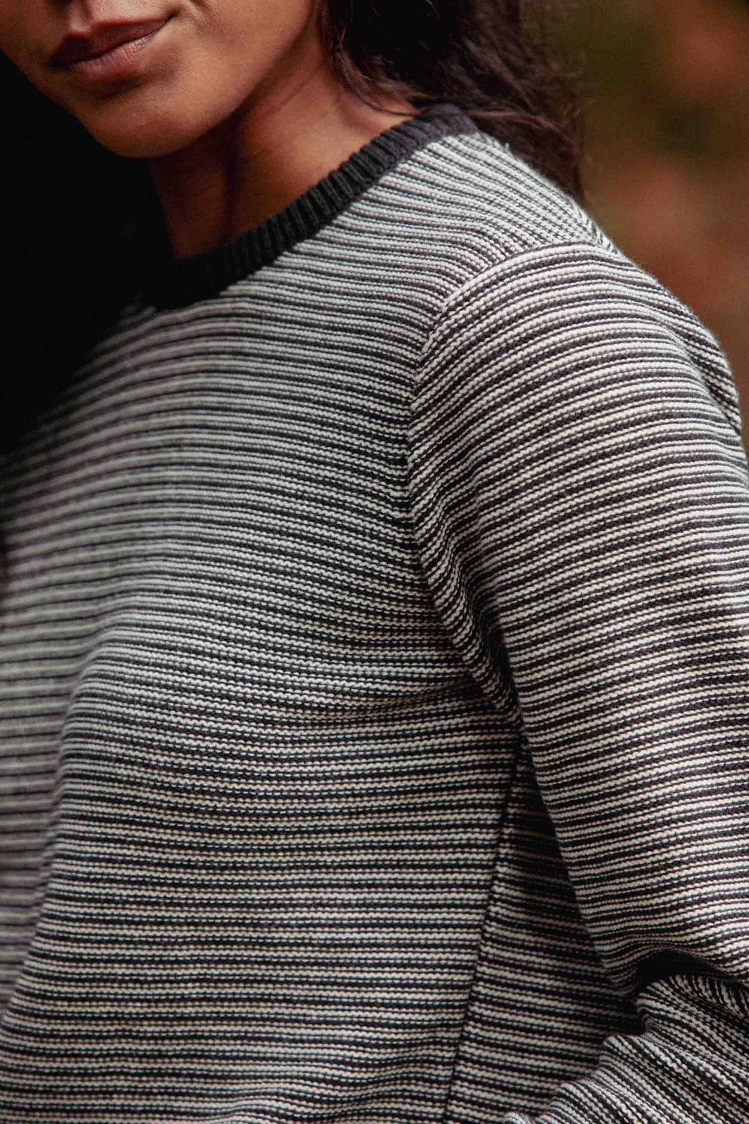 A close-up of a person outdoors wearing the Bridge & Burn Ansel Sweater in Charcoal Stripe, a mid-weight piece crafted from recycled fibers, with part of their face and upper body visible.