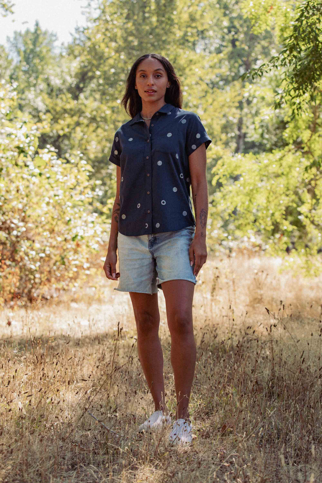 Standing outdoors on dry grass with trees behind, a person wears Bridge & Burn’s Bea Shirt in the Lunar Polka pattern, paired with light denim shorts and white sneakers.
