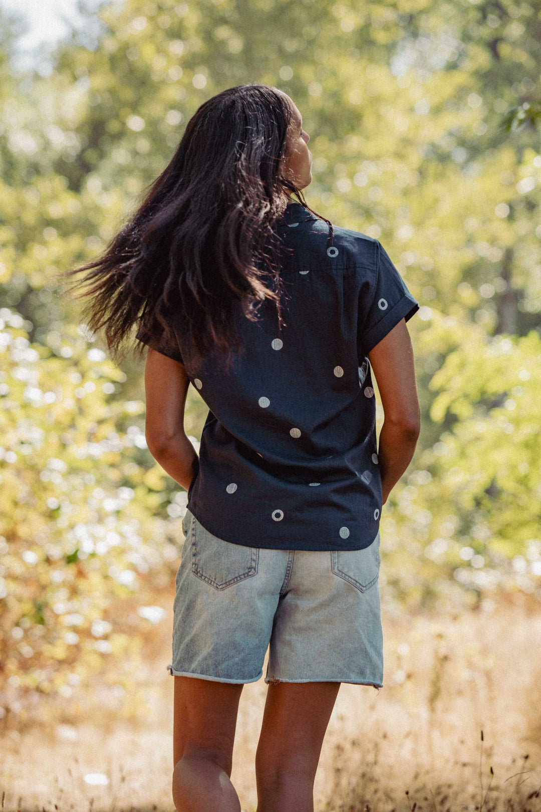 A person with long dark hair, wearing the Bridge & Burn Bea Shirt in Lunar Polka and denim shorts, stands outdoors with hands in pockets, surrounded by greenery and sunlight.
