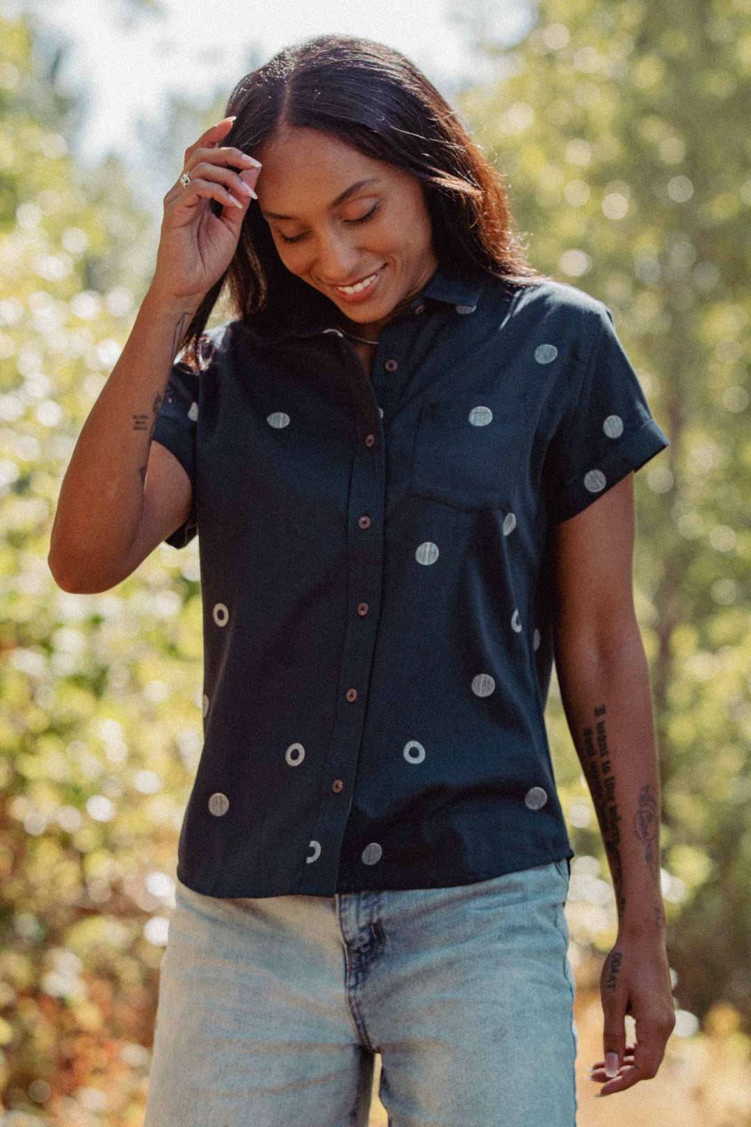 A person with long dark hair smiles and looks down while standing outdoors, wearing Bridge & Burn's Bea Shirt in Lunar Polka and light blue jeans. Sunlight filters through green trees behind them.