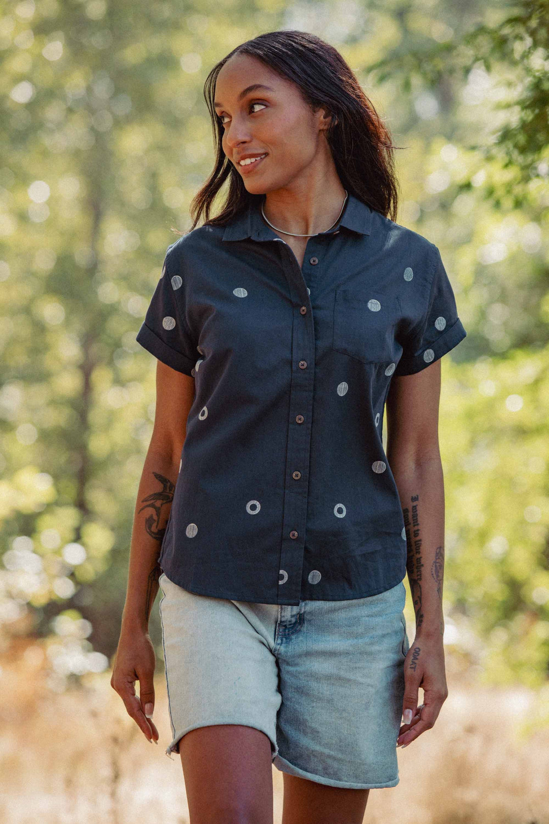 A woman with long hair wears the Bridge & Burn Bea Shirt in Lunar Polka and light denim shorts. She stands outdoors, smiling amid sunlight and lush green foliage.