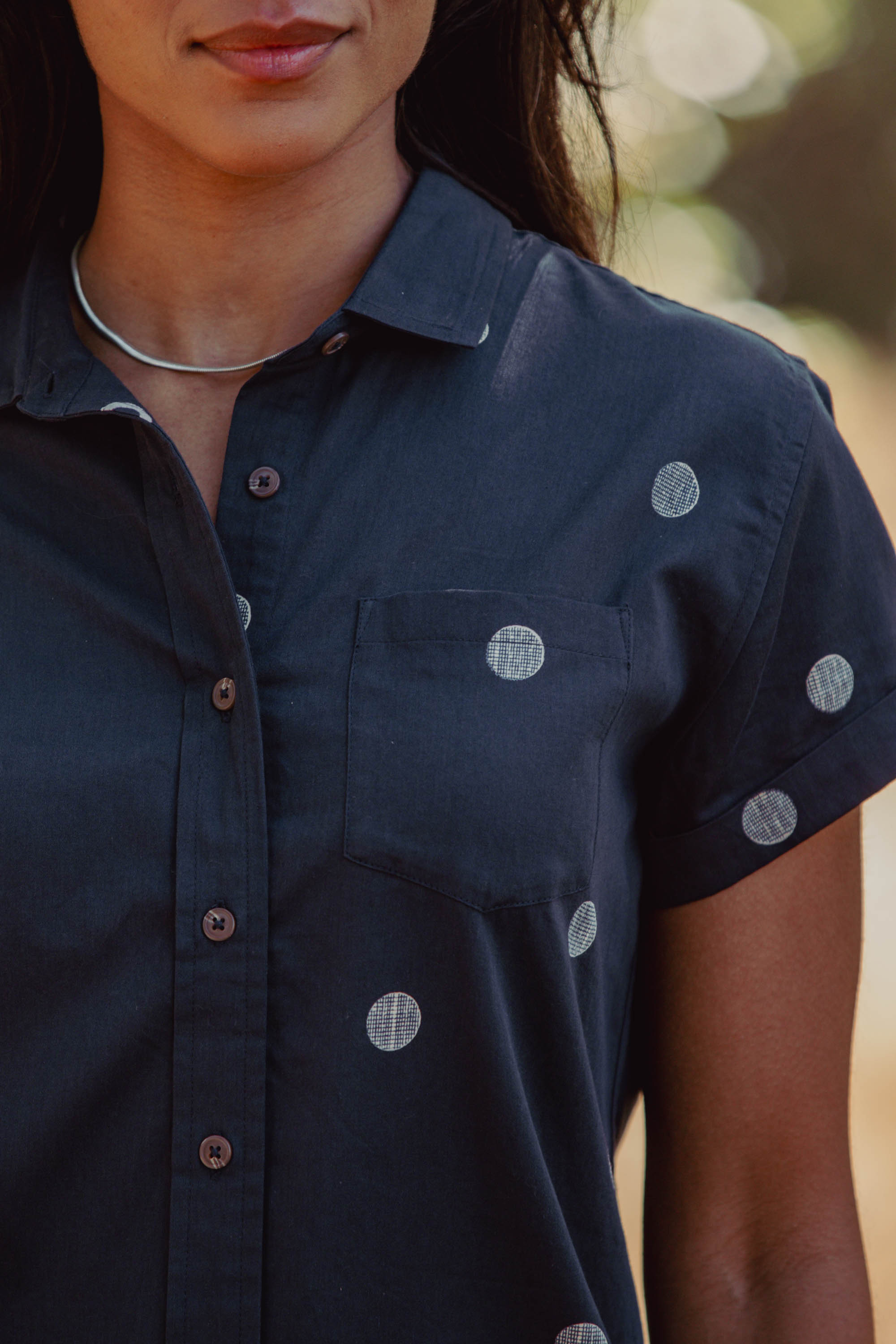 A woman wears the Bridge & Burn Bea Shirt in Lunar Polka, a dark tailored organic cotton short-sleeve button down with white polka dots and a silver necklace, photographed from shoulders to mid-torso against an outdoor, blurred background.