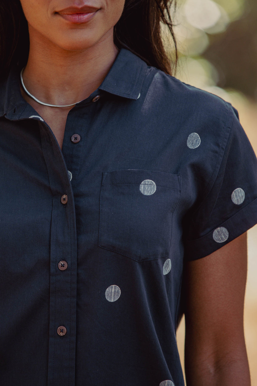 A woman wears the Bridge & Burn Bea Shirt in Lunar Polka, a dark tailored organic cotton short-sleeve button down with white polka dots and a silver necklace, photographed from shoulders to mid-torso against an outdoor, blurred background.