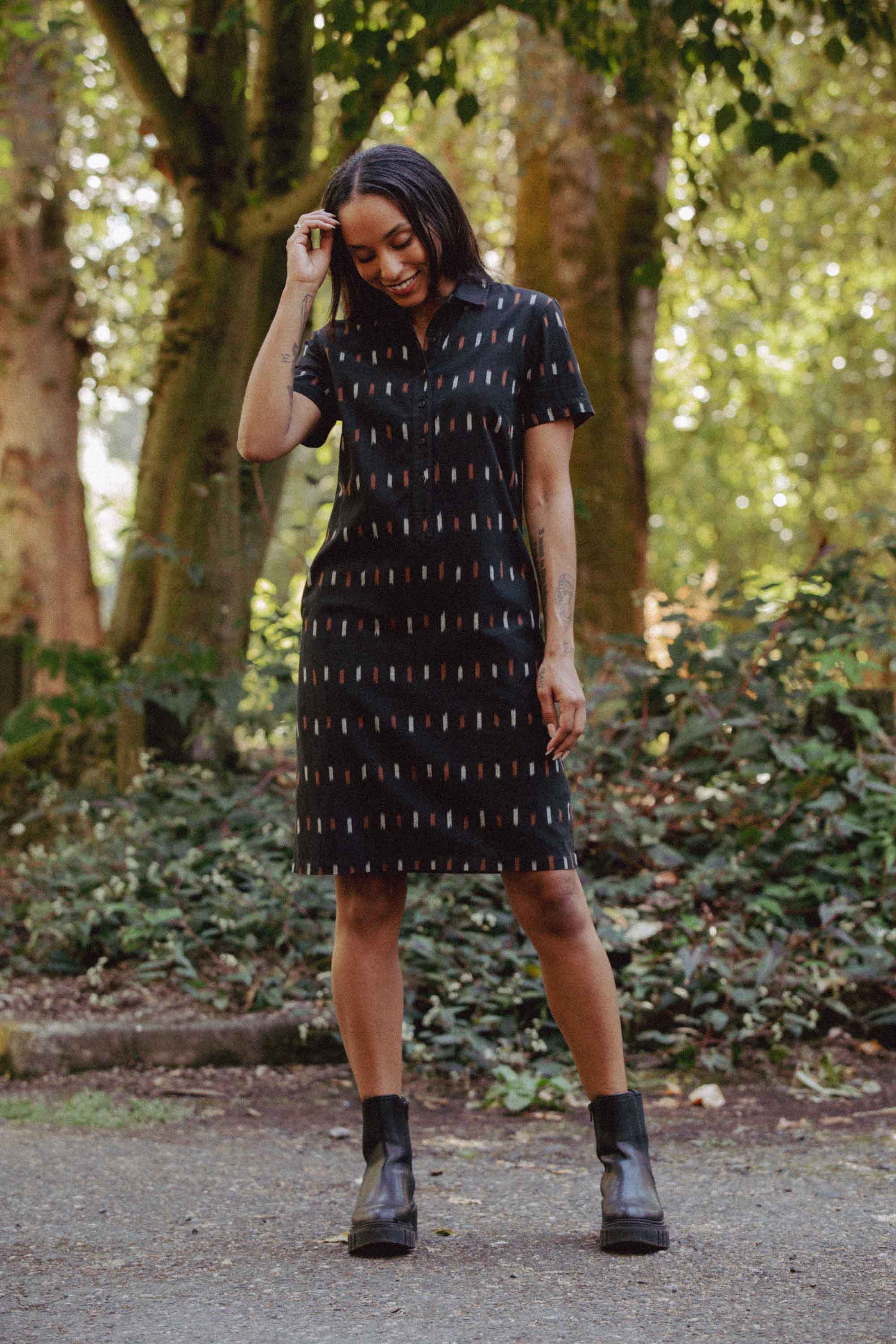 A woman smiles outdoors on a path, wearing the Bridge & Burn Delia Dress in Dotted Horizon—a short-sleeved organic cotton shirt dress with a colorful pattern—and black ankle boots, surrounded by trees and greenery.