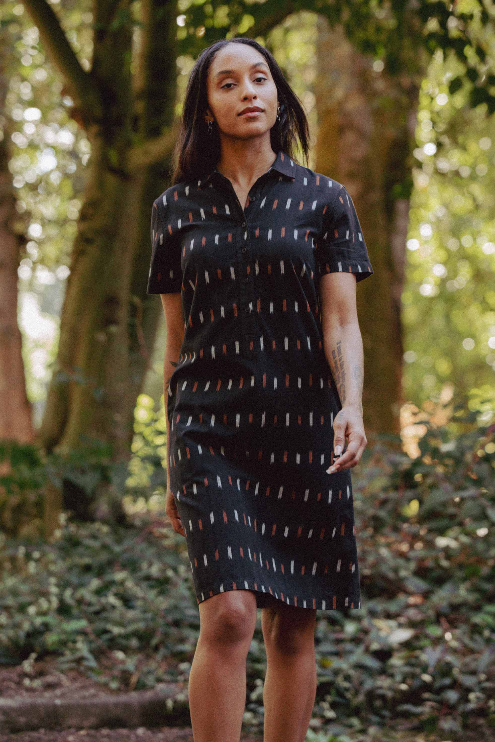 A woman strolls through the woods in the Bridge & Burn Delia Dress / Dotted Horizon, a short-sleeved, knee-length black shirt dress with colorful dashes, made from organic cotton. The dress suits her long dark hair and serene look.