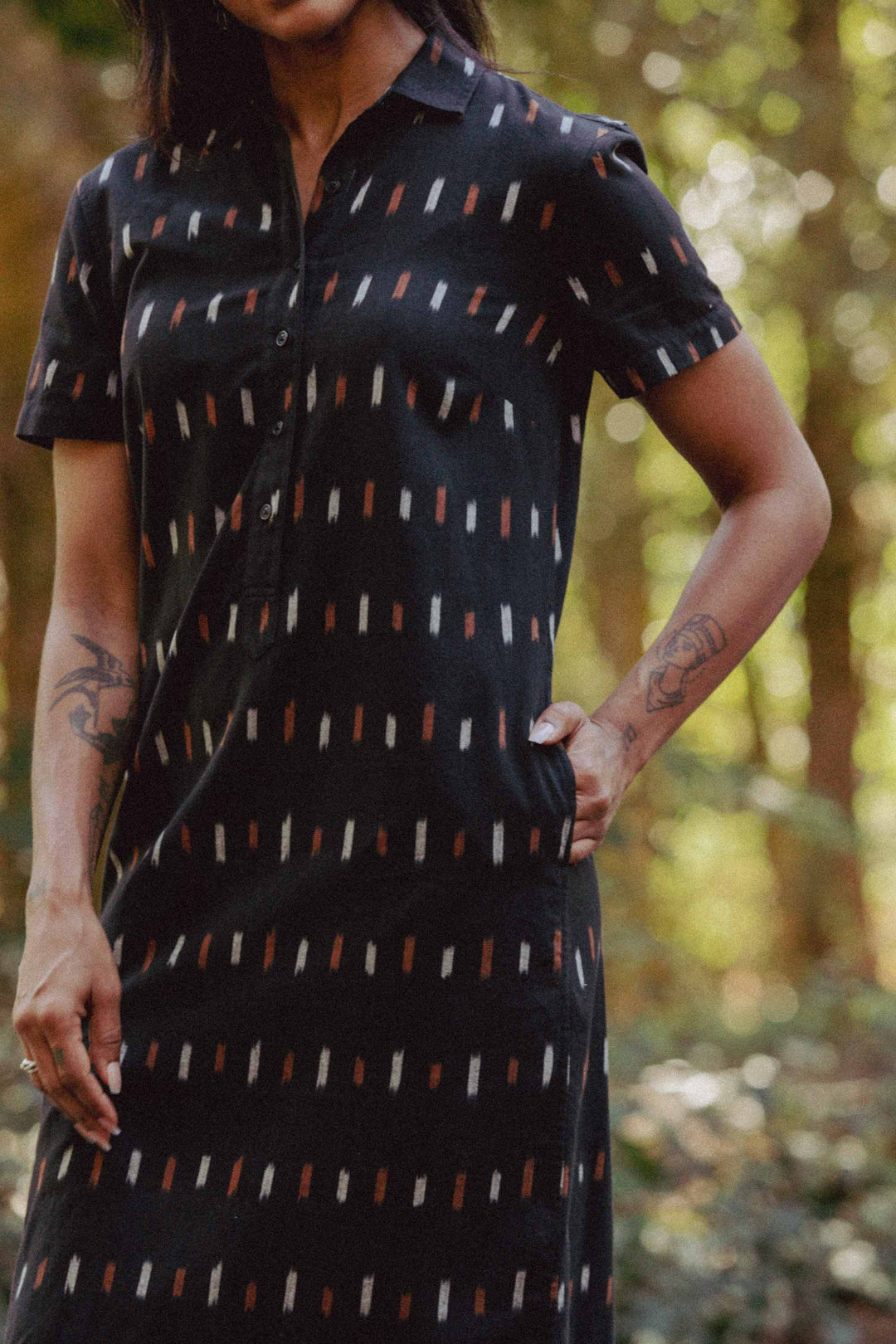 A person stands outdoors, sunlight and trees behind them, wearing Bridge & Burn's Delia Dress in Dotted Horizon—a short-sleeved black shirt dress with a vertical pattern. Their left hand is in their pocket; tattoos are visible on both arms.