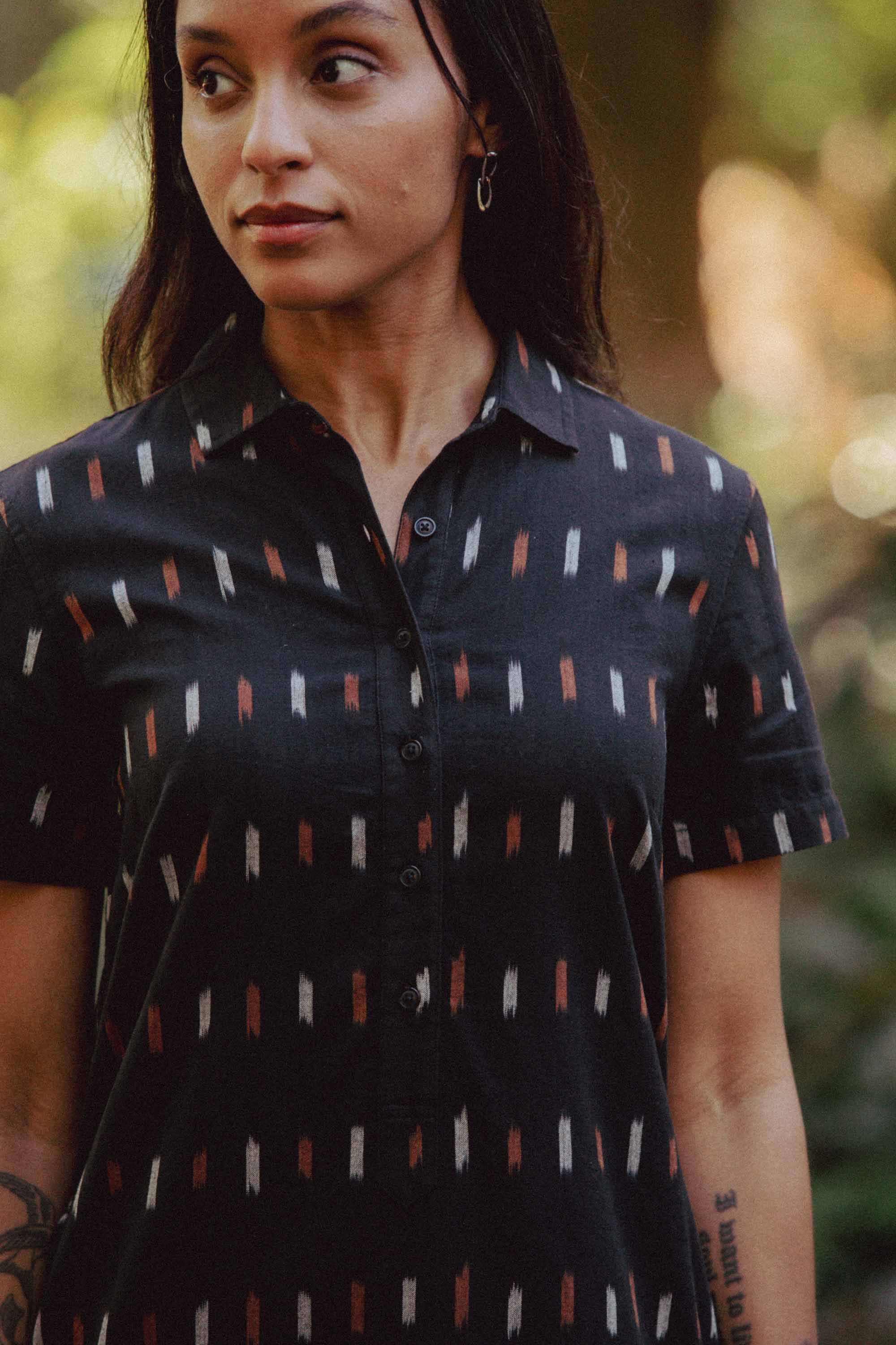 A woman with long dark hair and a forearm tattoo stands outdoors, wearing the Bridge & Burn Delia Dress in Dotted Horizon, featuring short sleeves and vertical white, brown, and rust dashes. She looks slightly to the side.