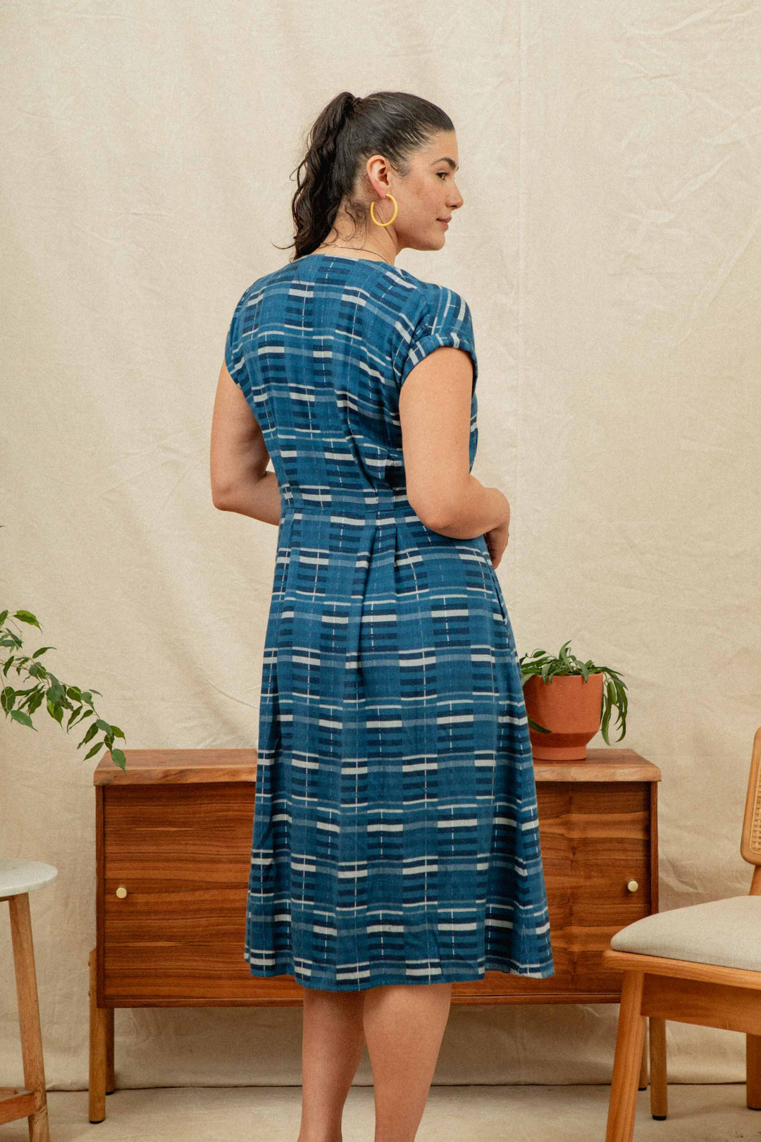A woman with dark hair in a ponytail stands with her back to the camera, wearing the Eileen Dress in Stormcrest Stripe by Bridge & Burn, in a room with wooden furniture and potted plants.