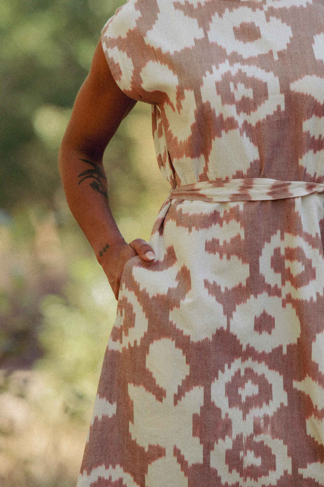 A person stands outdoors with one hand in the pocket of the Eloise Dress / Pop Leaf by Bridge & Burn. The 100% cotton, abstract-patterned A-line dress highlights a tattooed forearm against a backdrop of blurred greenery.