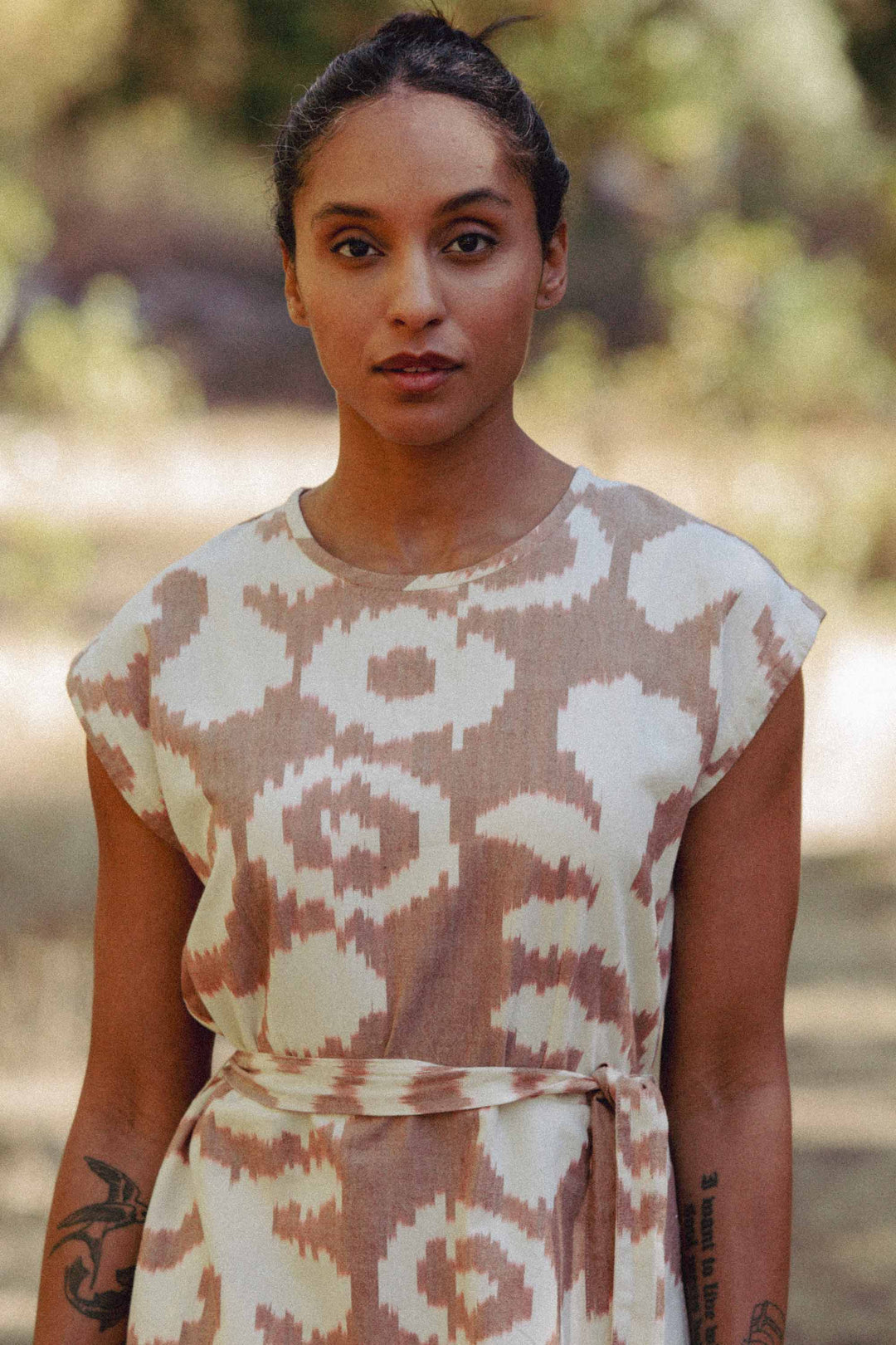 A woman with dark hair tied back stands outdoors amid blurred greenery, wearing the Bridge & Burn Eloise Dress in Pop Leaf—a short-sleeved beige and white A-line dress. She has tattoos on her left arm and a calm expression.