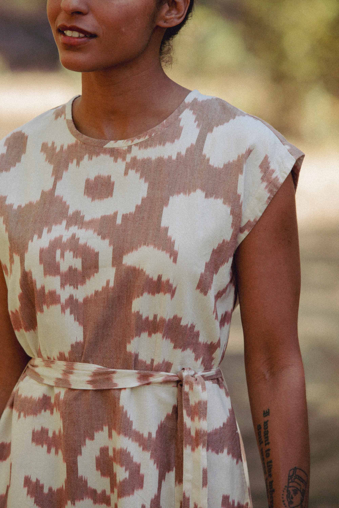 A person stands outdoors in the Eloise Dress / Pop Leaf by Bridge & Burn—a beige and white patterned, 100% cotton A-line dress with cap sleeves and matching waist tie. Their tattooed arm is visible, face partially shown with a soft smile.