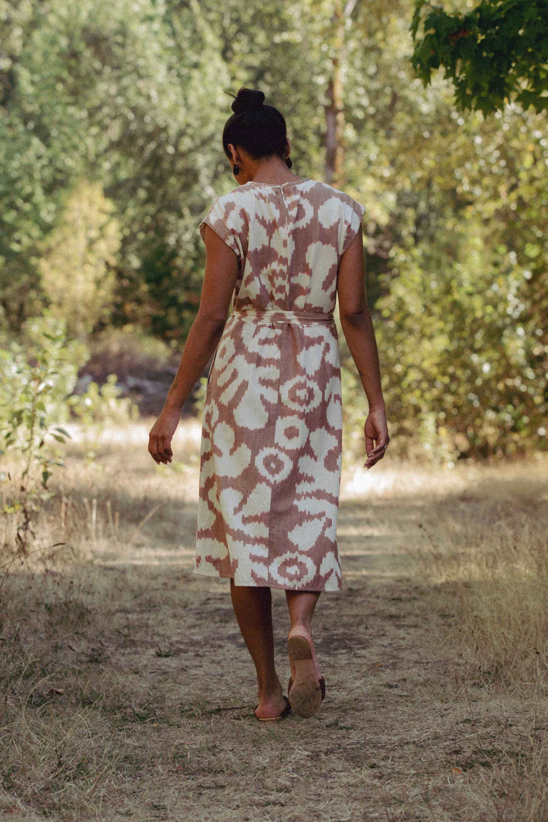 A person in the Eloise Dress / Pop Leaf by Bridge & Burn walks barefoot on a sunlit forest path, their back to the camera—capturing the timeless charm of this patterned A-line wardrobe essential.