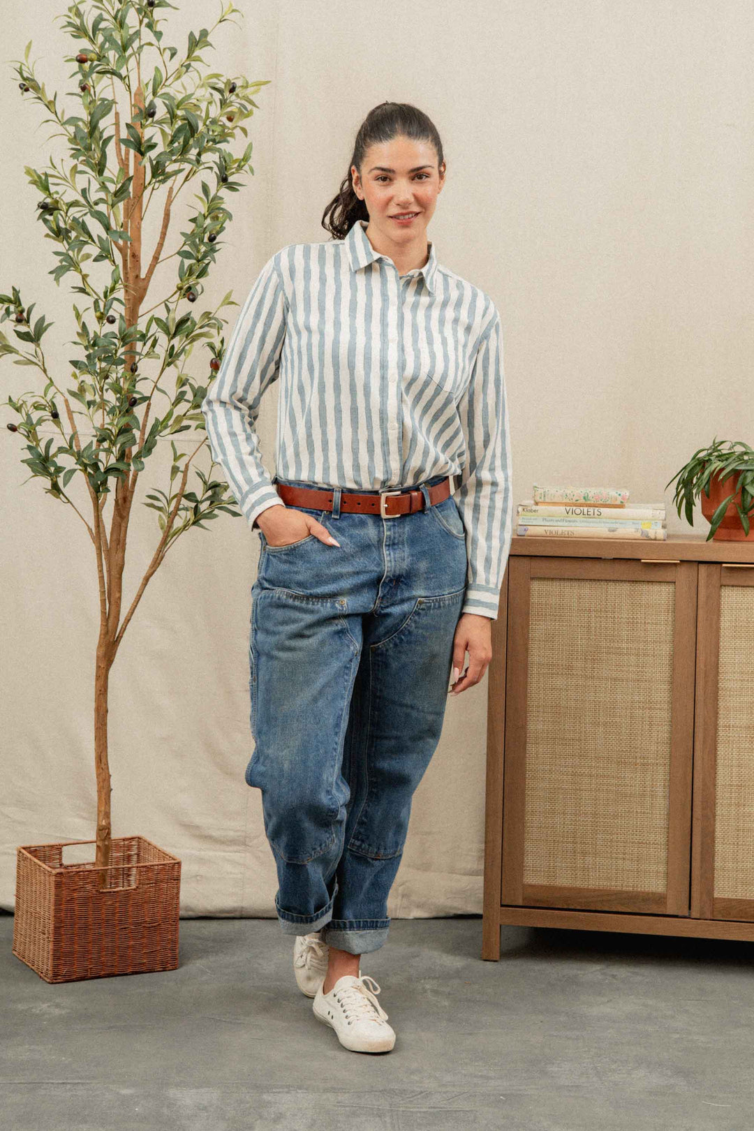A woman stands indoors next to a potted plant and cabinet, wearing the Bridge & Burn Haley Shirt in Beach Stripe with blue cuffed jeans, a brown belt, and white sneakers. She smiles gently, one hand in her pocket.