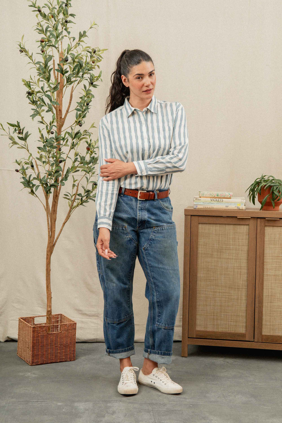 A woman with long dark hair in a ponytail stands indoors near a potted plant and cabinet, wearing the Bridge & Burn Haley Shirt in Beach Stripe with cuffed blue jeans, white sneakers, and a brown belt.