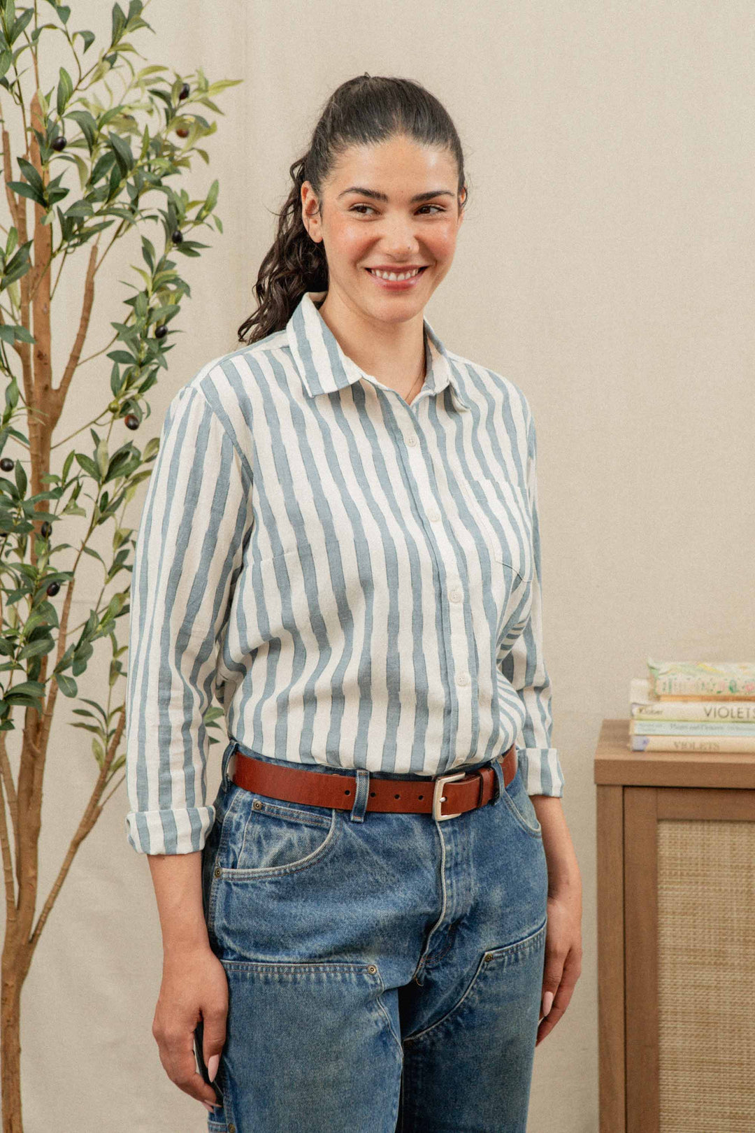 A woman with long dark hair in a ponytail smiles while wearing the Bridge & Burn Haley Shirt in Beach Stripe, styled with blue jeans and a brown belt. She stands indoors near a leafy plant and a wooden cabinet with books on top.