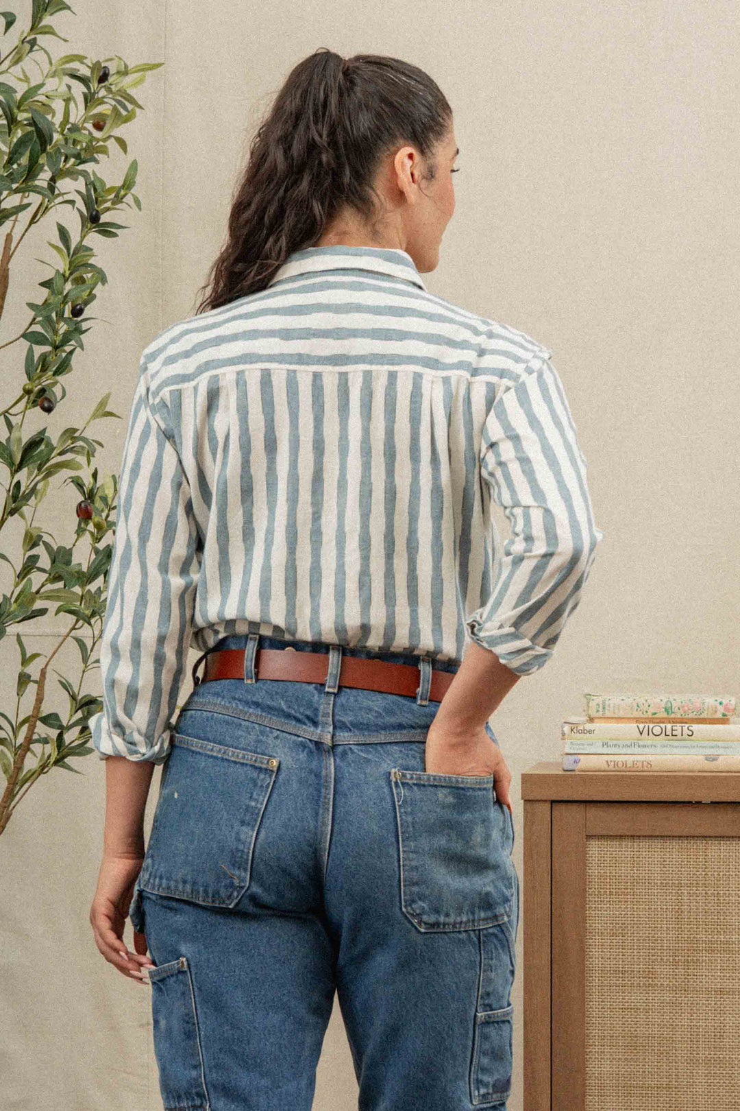 A person with long dark hair in a ponytail wears the Bridge & Burn Haley Shirt in Beach Stripe, paired with blue jeans and a brown belt, standing with their back to the camera near a plant and books on a cabinet.