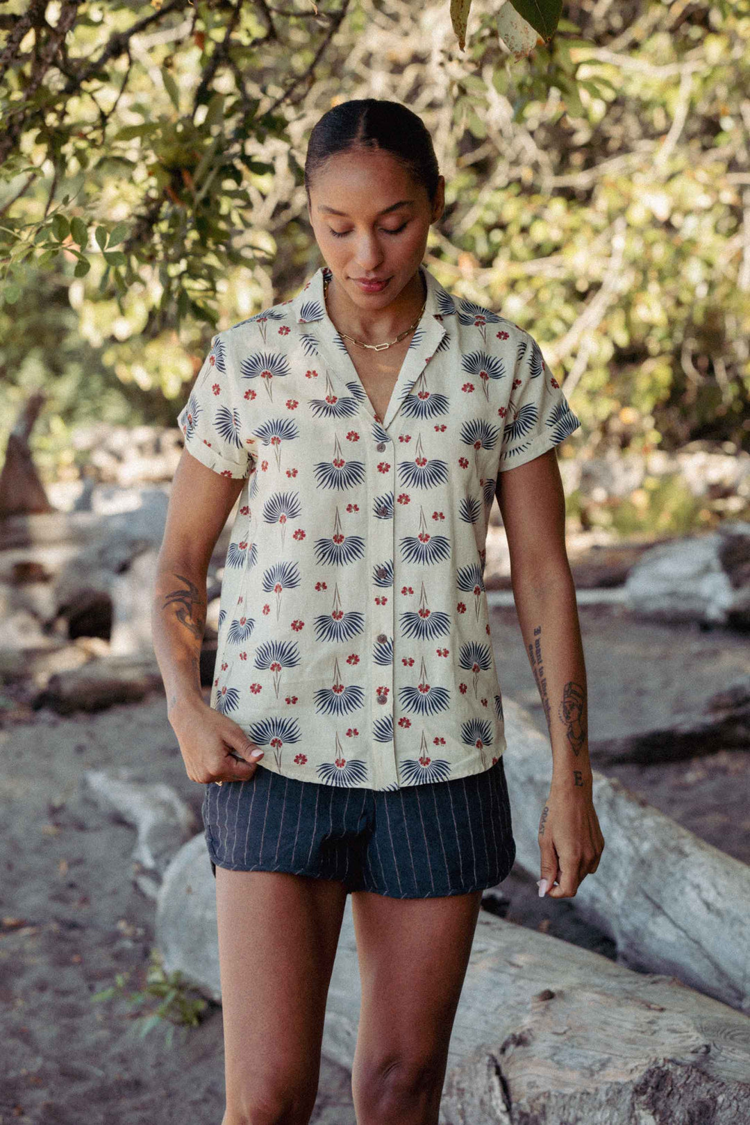 A person stands outdoors on a sandy, wooded area wearing the Bridge & Burn Innes Shirt in Aloha Floral. They look down with hands in pockets as sunlight filters through the trees in the background.