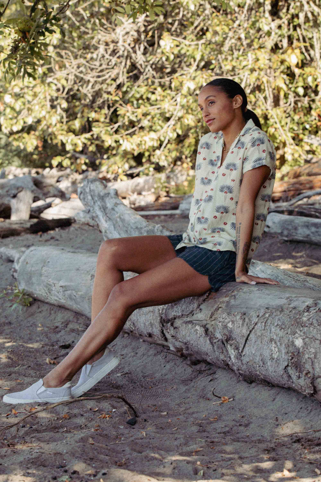 A woman sits on driftwood outdoors, wearing the Bridge & Burn Innes Shirt in Aloha Floral, dark pinstripe shorts, and white slip-on sneakers, with trees and sandy ground in the background.