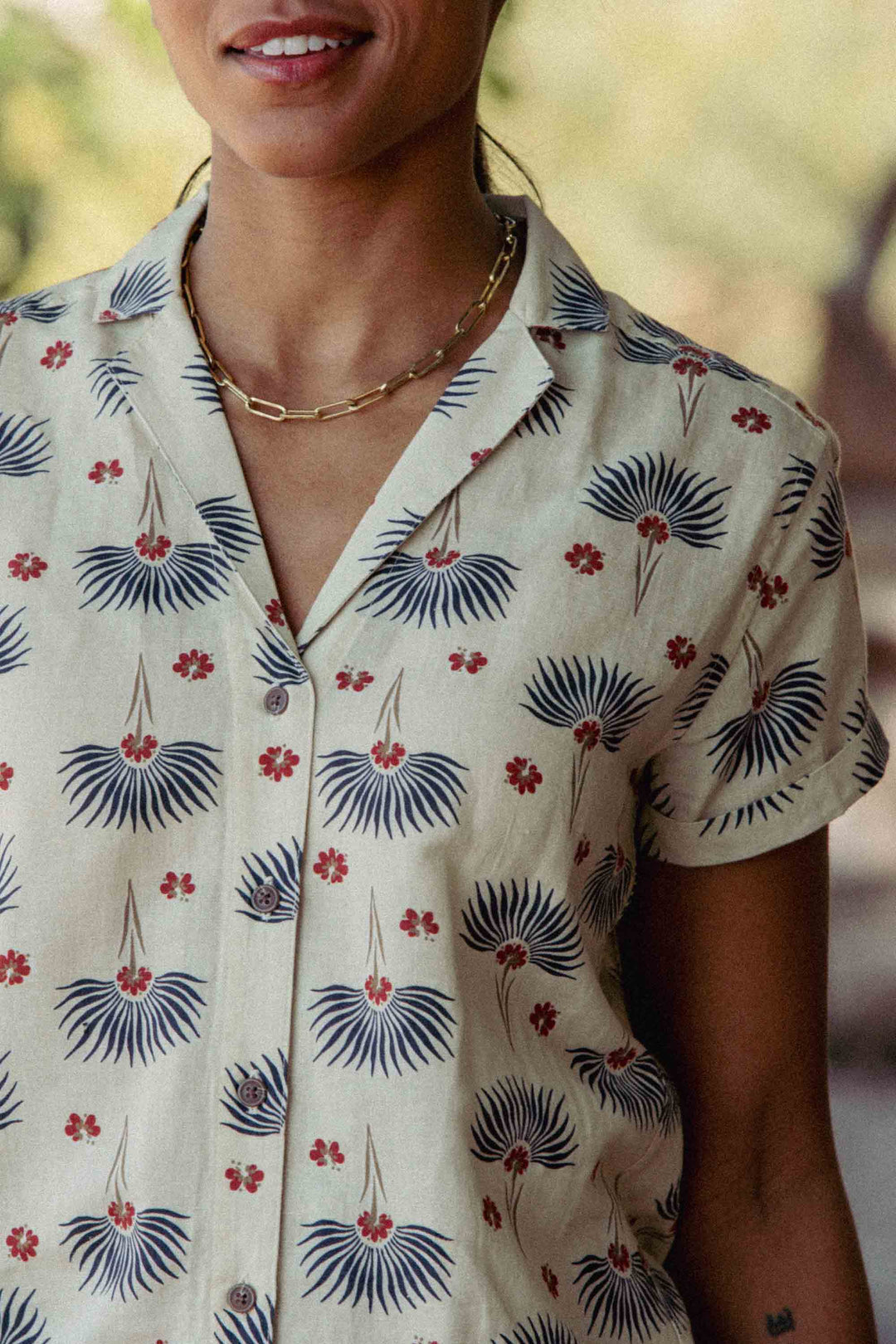 A person wears the Bridge & Burn Innes Shirt in Aloha Floral—a relaxed fit button-up with blue fan-shaped designs and red flowers—paired with a gold chain necklace. They are smiling, and their face is partially visible.