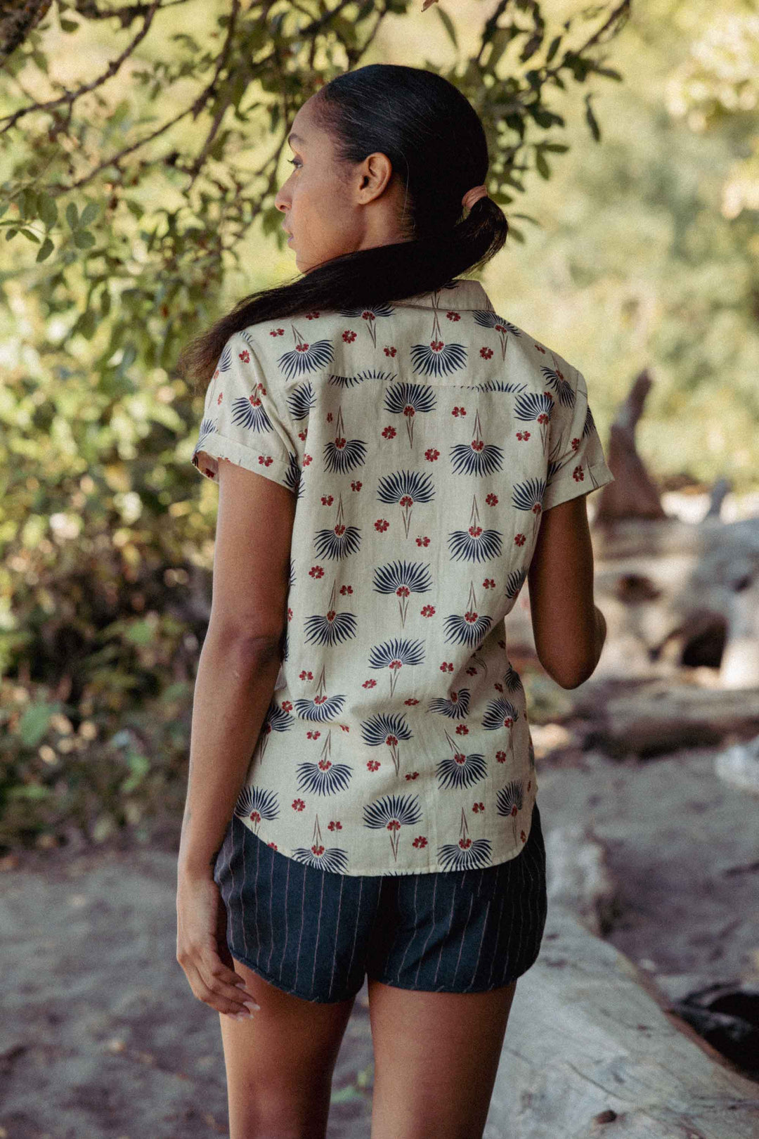 A woman stands outdoors with her back to the camera, wearing the Bridge & Burn Innes Shirt in Aloha Floral and dark striped shorts, near greenery and fallen logs.